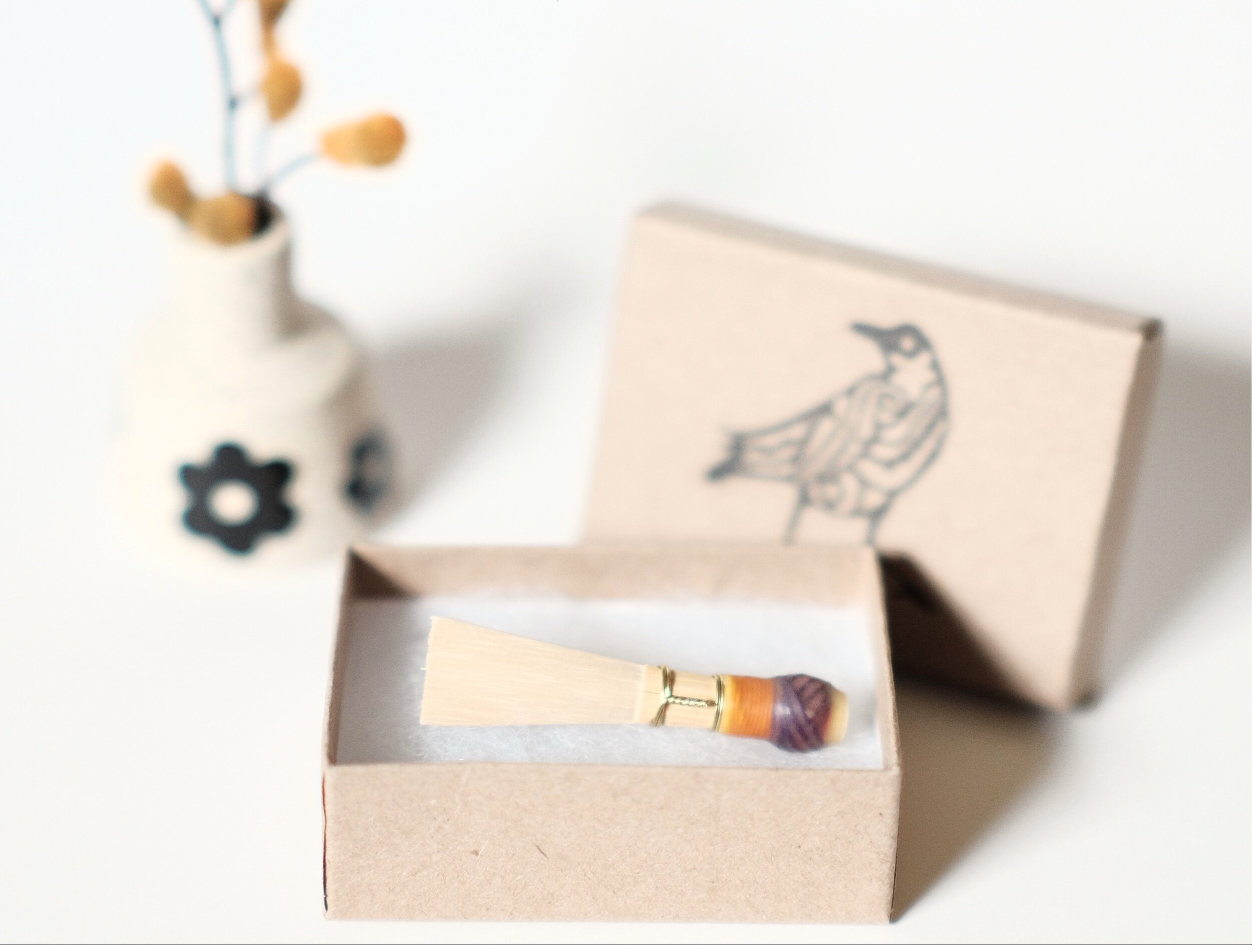 One bassoon reed blank sits in a cardboard jewelry box showing the top with the Raven Reeds logo stamped and a small vase with orange flowers in the background