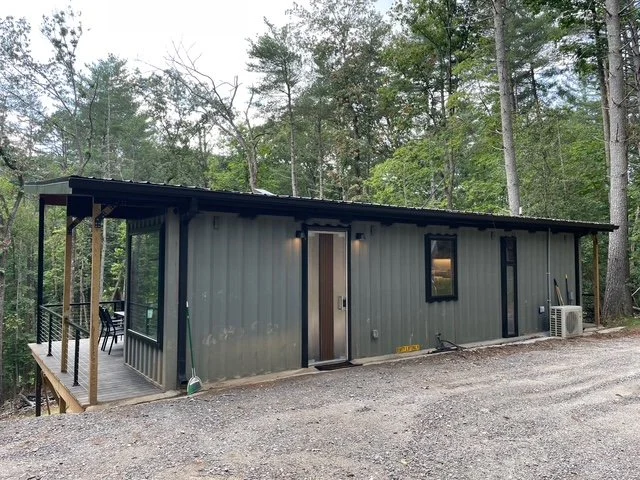 Shipping Container Home with metal roof, surrounded by trees, featuring a front porch with a railing and outdoor seating.