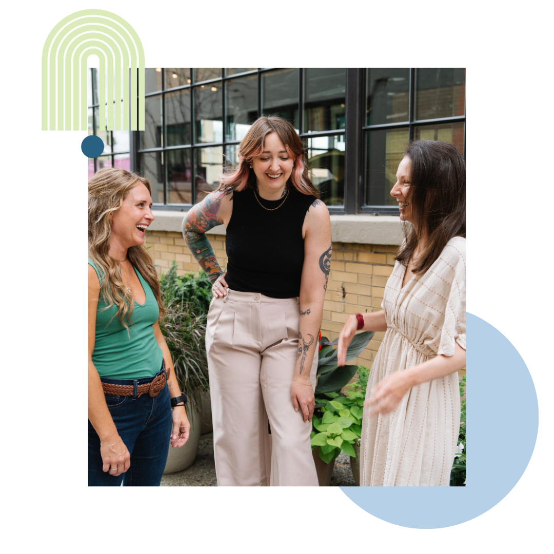 Three women on the Green Loop team, one with long blonde hair, one with brown and pink hair, and one with long dark brown hair, have a conversation and laugh outdoors.