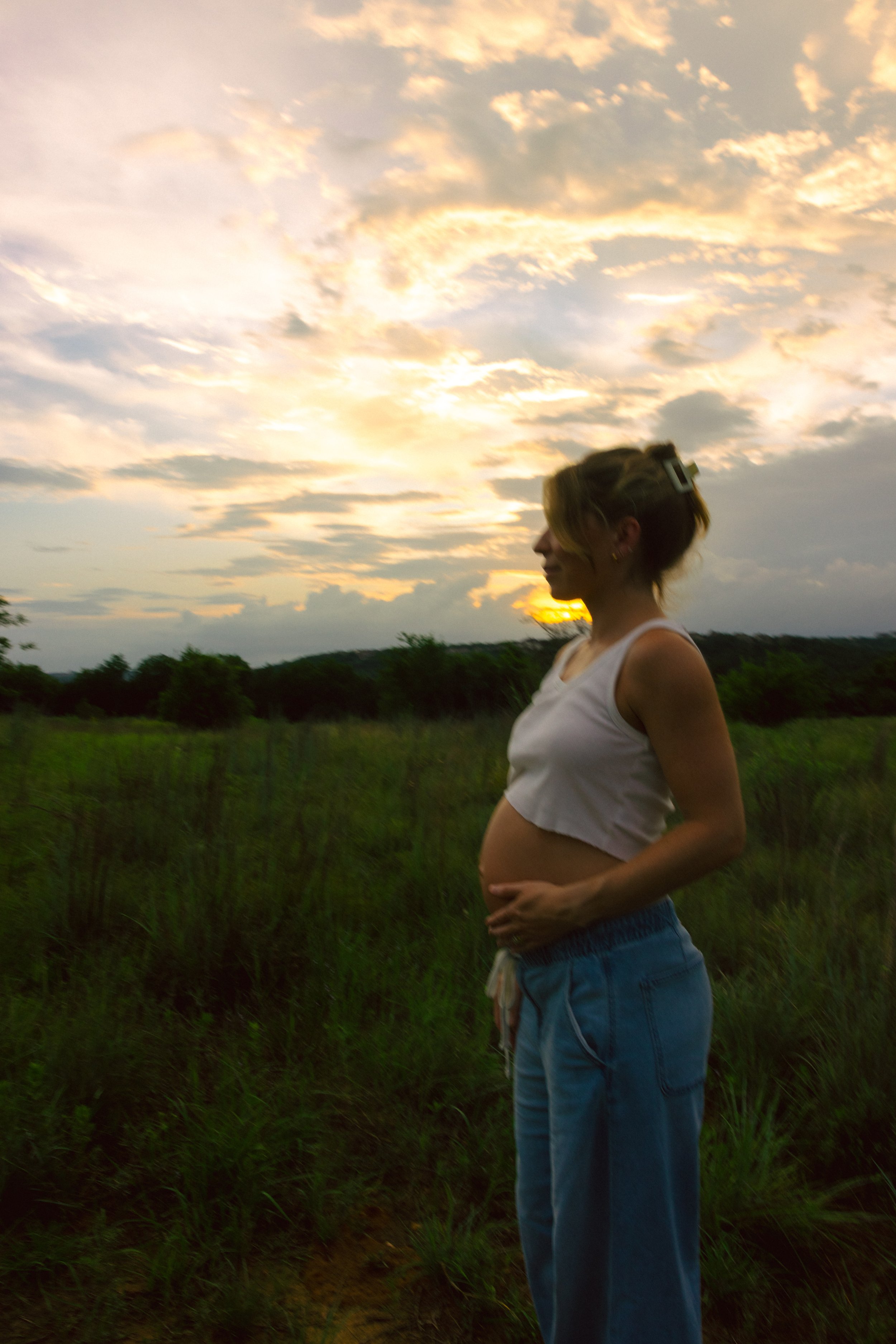 A pregnant woman with short hair and earrings standing outdoors at sunset, wearing a sleeveless top and jeans, with her hands on her belly, in a green field with trees and a cloudy sky in the background.