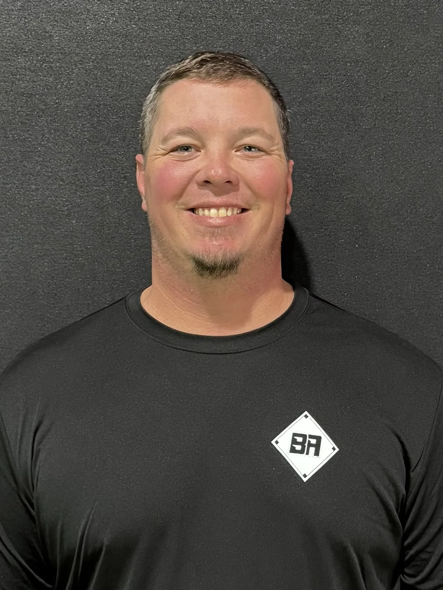 Smiling person wearing a black shirt with "BA" logo, against a dark background.