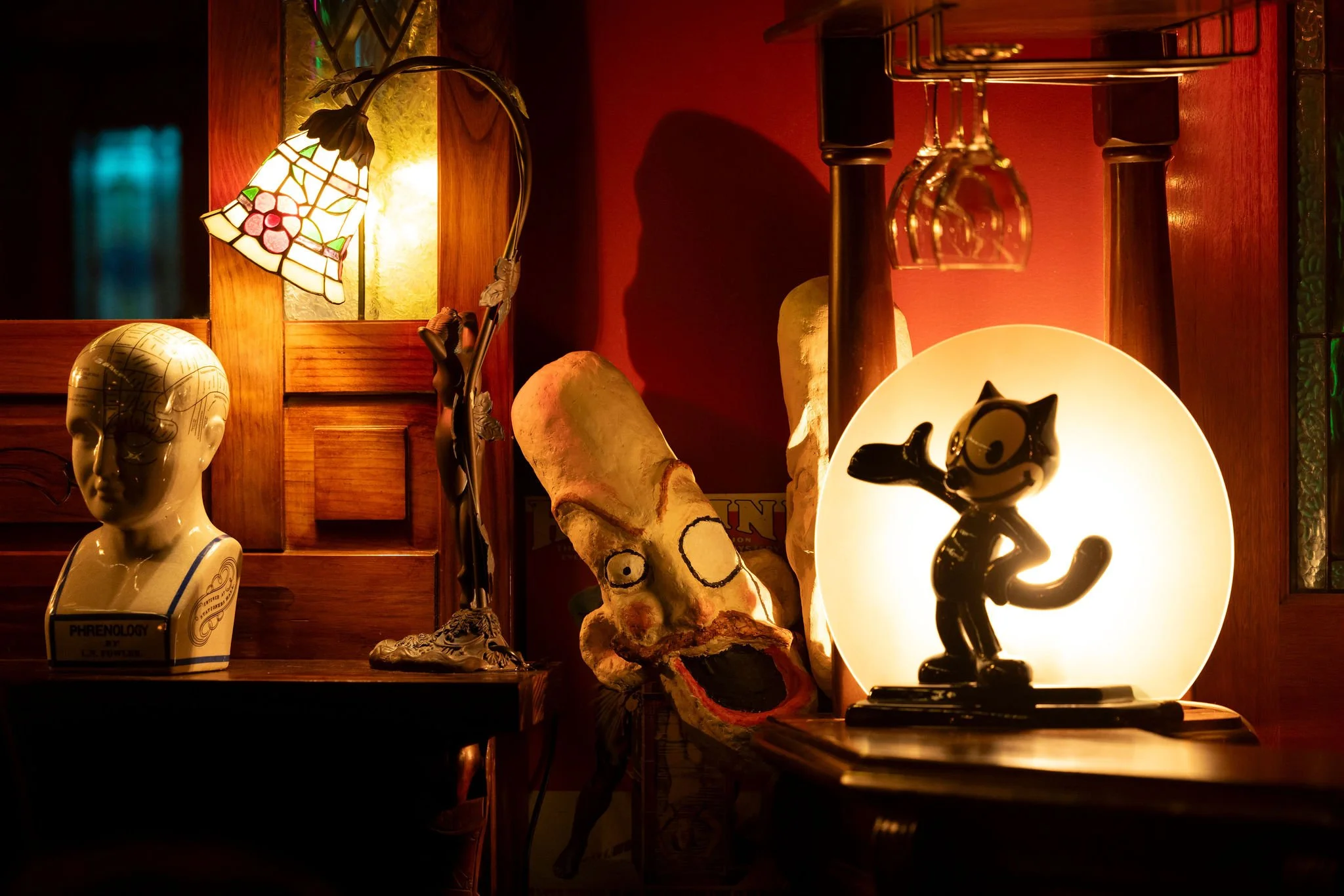 A dimly lit wooden shelf with eclectic decorations including a ceramic head, a lamp with a stained-glass lampshade depicting grapes, a carved animal figure, a skull with glasses, and various decorative objects.