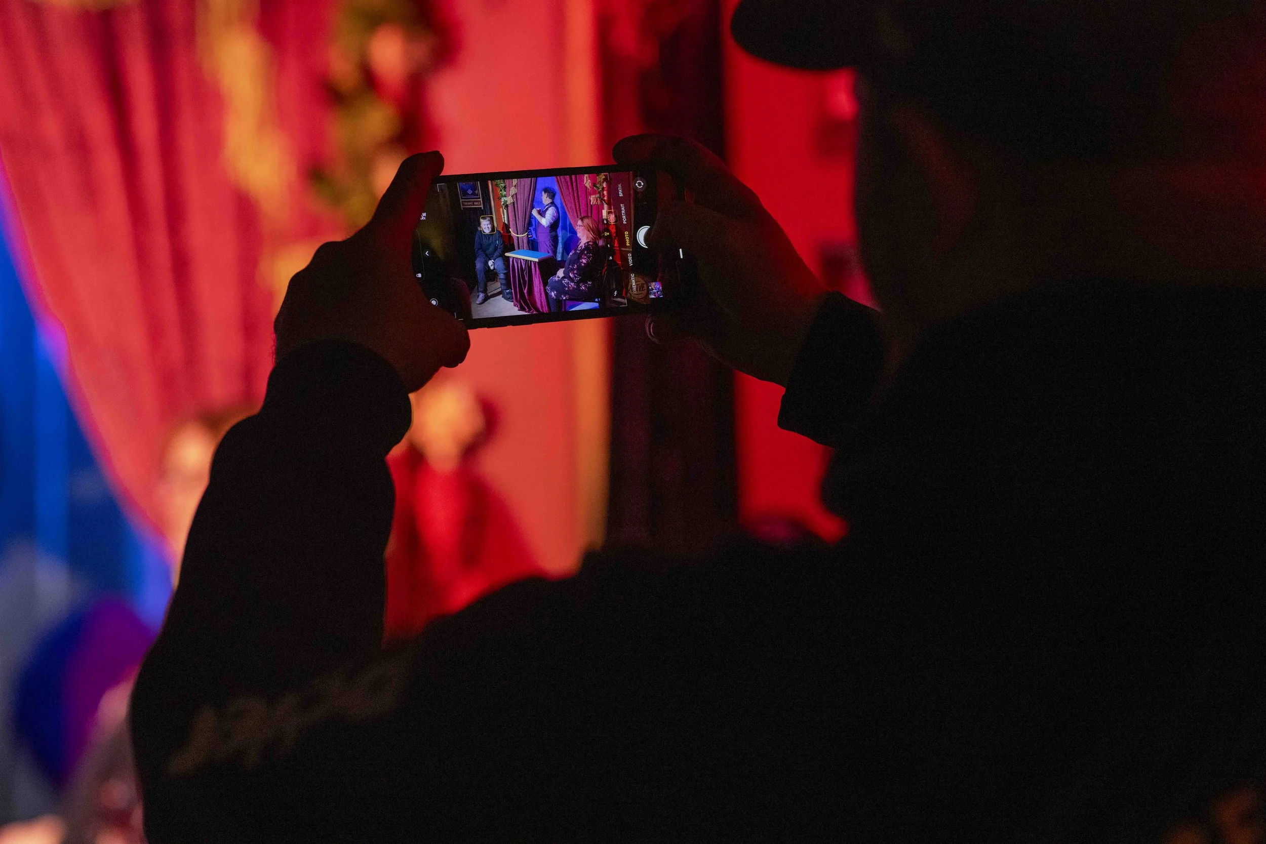 Person taking a photo or recording a video with a smartphone in a dimly lit environment with red and blue lighting, showing a scene on the phone where two people are seated on a stage with red velvet curtains and purple drapes.