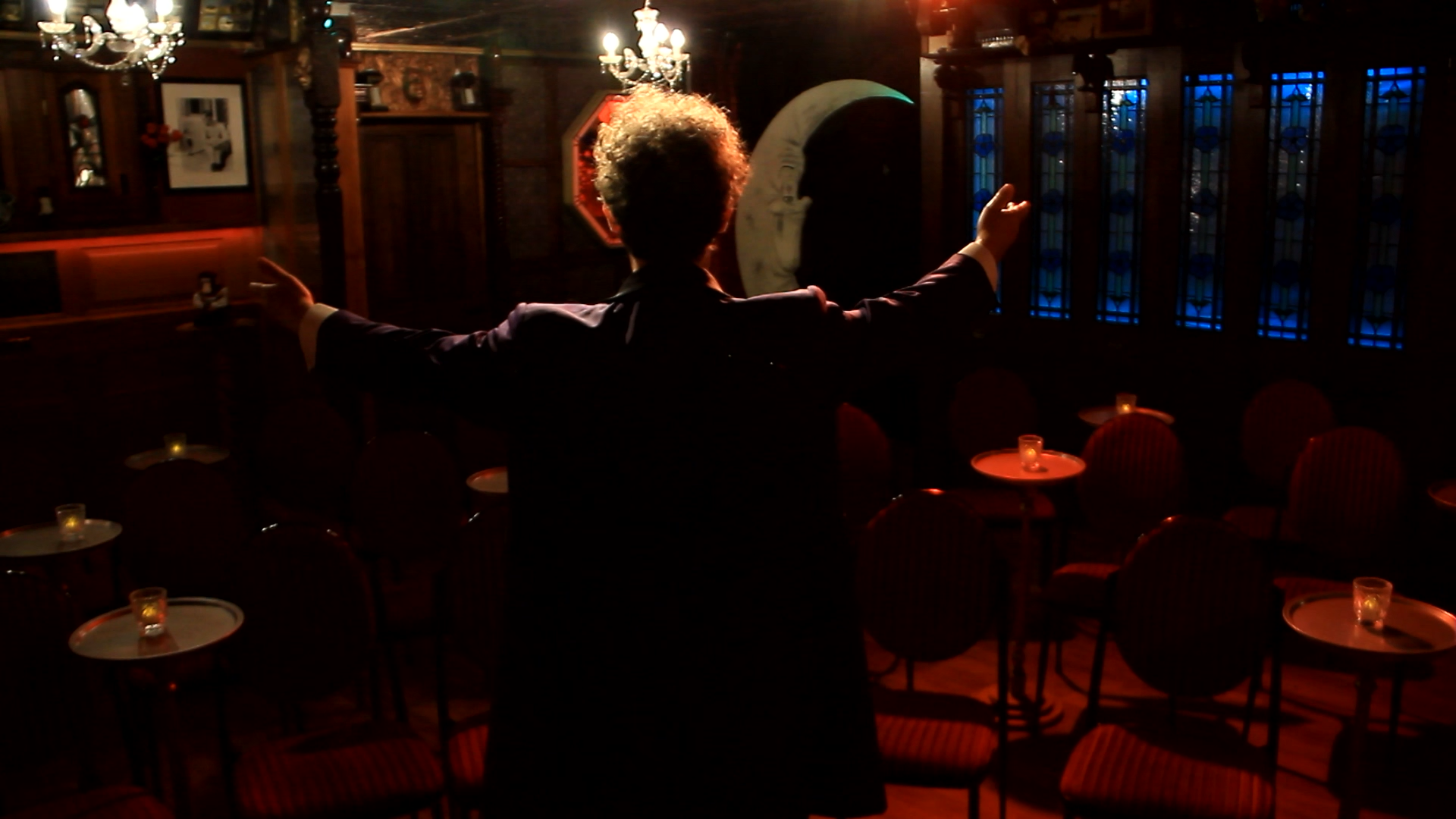 A person with curly hair, wearing a dark suit, stands with arms outstretched in an empty, dimly-lit restaurant or bar with rows of small tables and chairs, chandeliers, and stained glass windows.