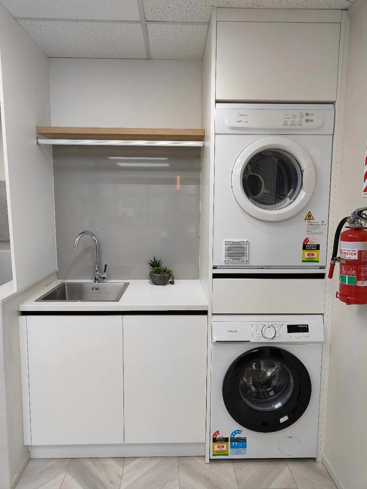 Customisable compact laundry cabinet with stacked washer and dryer, pull-out laundry hamper, integrated ironing board, sink, and overhead storage.