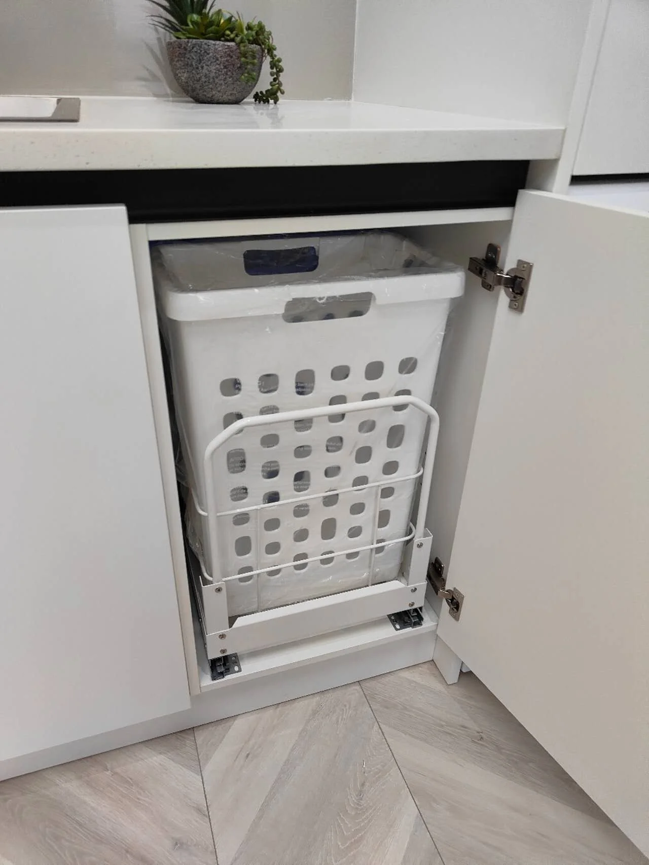 Pull-out laundry hamper installed inside a white laundry cabinet with removable basket and soft-close runners.