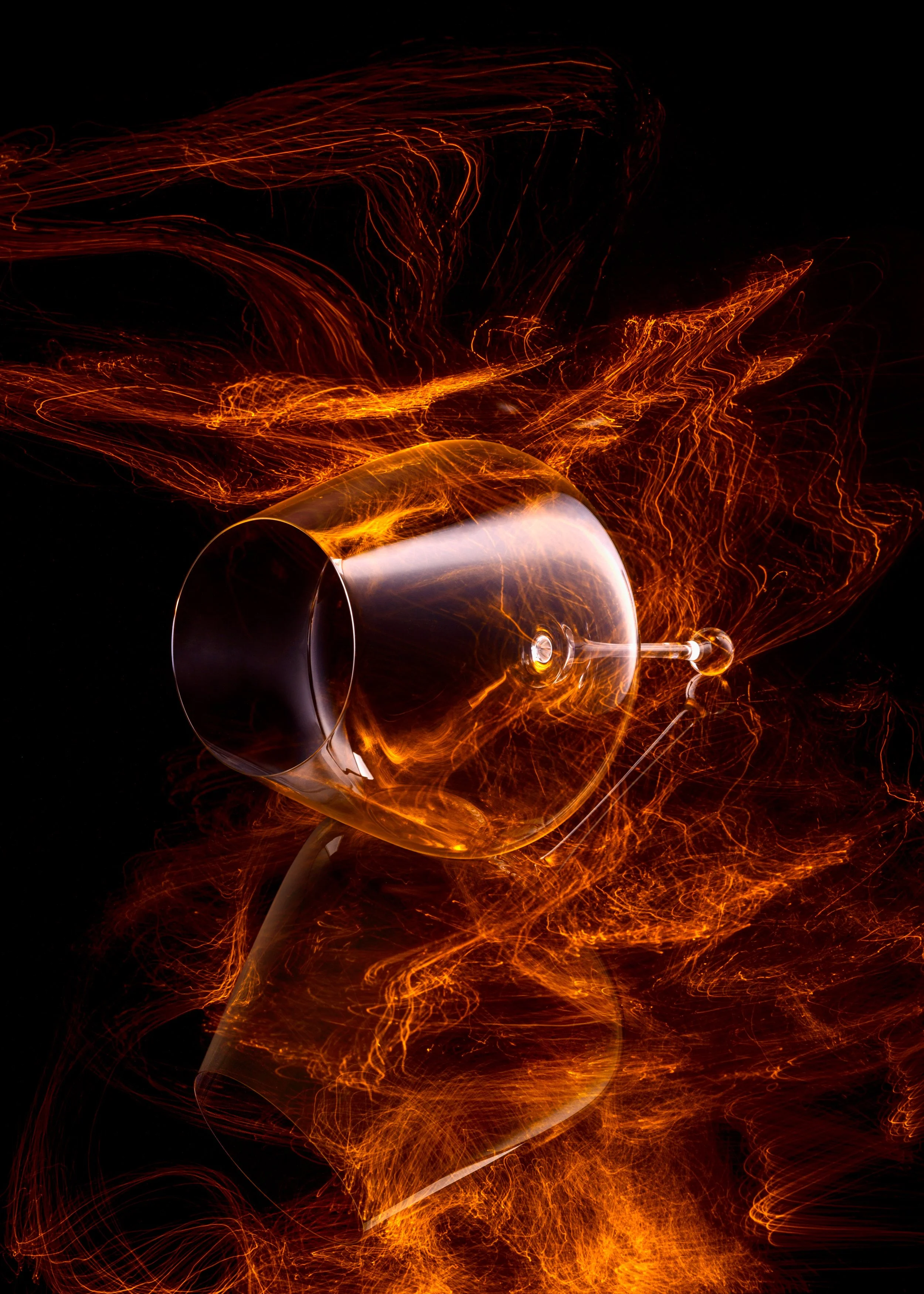 A wine glass lies on its side surrounded by dynamic, fiery streaks of orange light, creating an abstract composition against a black background.