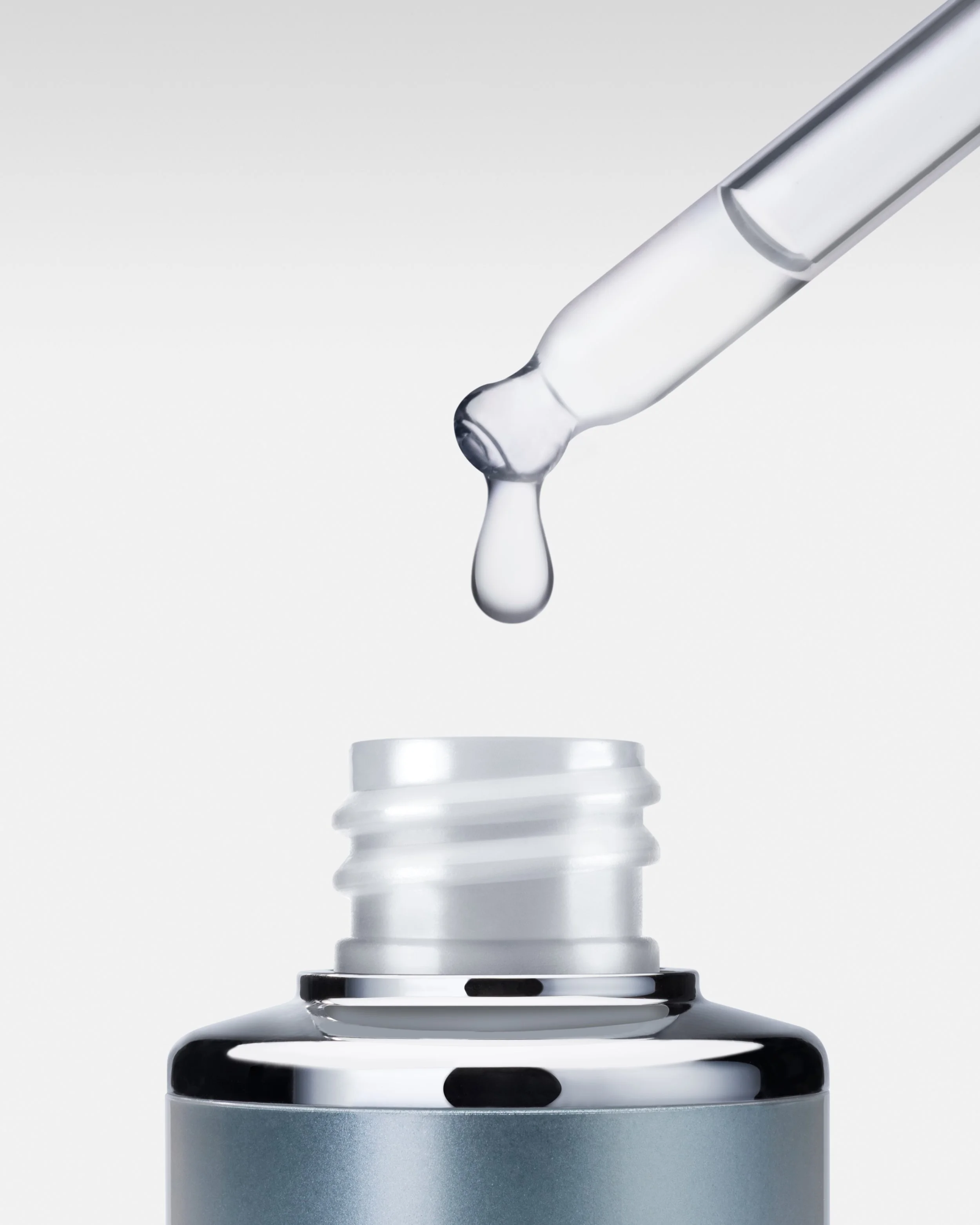 Close-up of a laboratory dropper releasing a clear liquid onto a layered glass platform with black and silver components, against a white background.