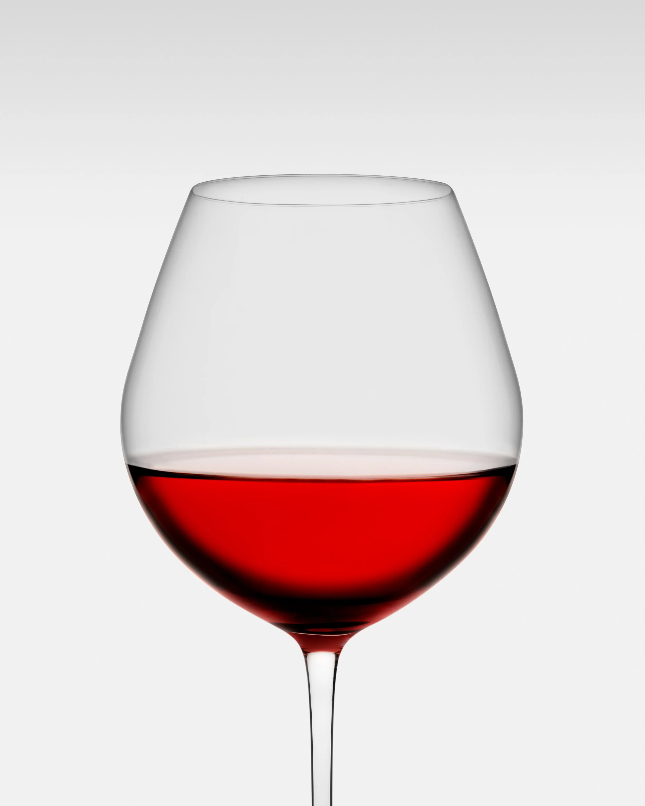 Modern stemmed wine glass filled with red wine, placed against a plain light background.