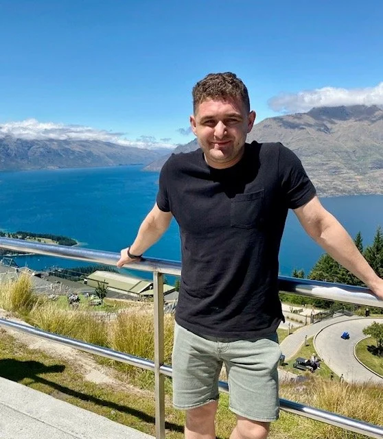 Man standing outdoors on a railing overlooking a lake and mountains, smiling, wearing a black t-shirt and gray shorts.
