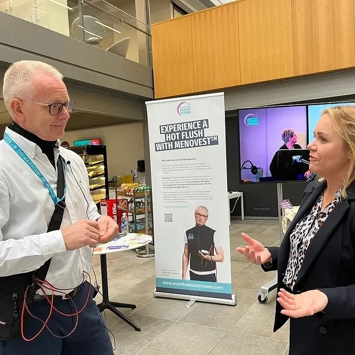Two people having a conversation in a modern indoor space with a promotional banner and TV screens in the background.