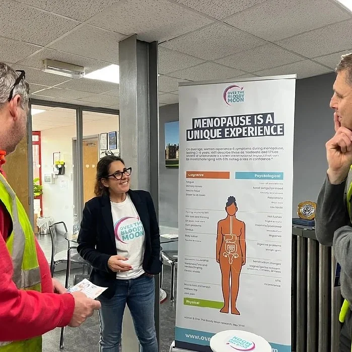 A woman is giving a presentation about menopause to two men in an indoor setting. The woman is wearing glasses and a shirt with the logo 'Over the Bloody Moon.' The presentation board highlights information about menopause, including cognitive, psychological, and physical aspects.
