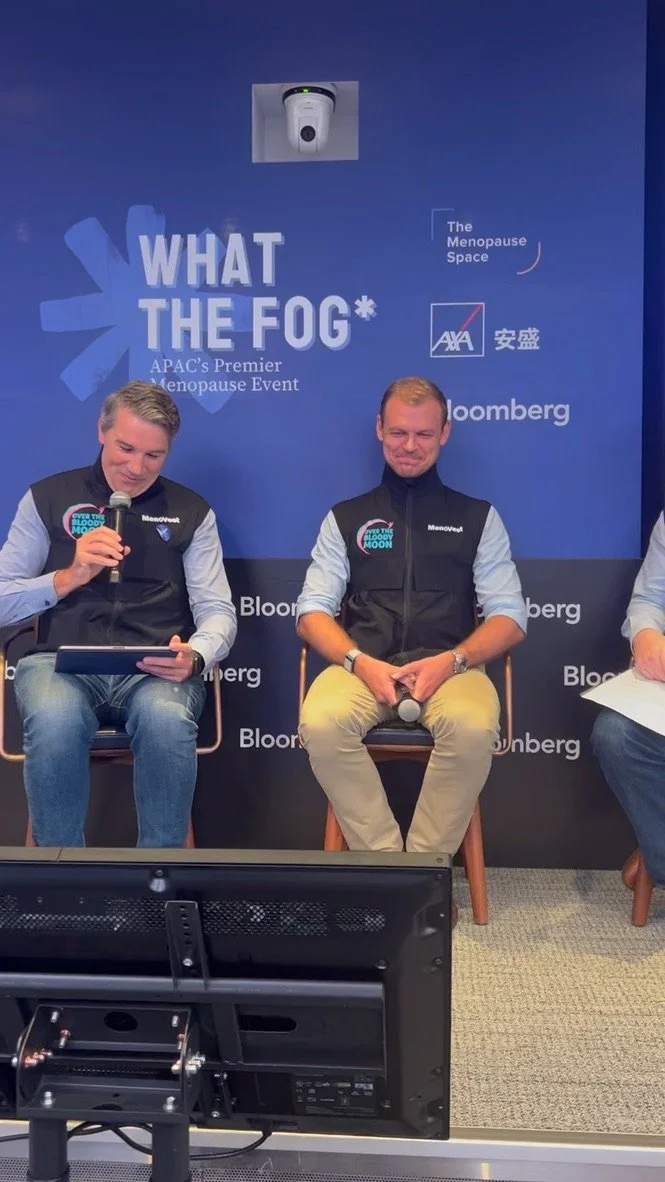 Panel discussion at a menopause event with a blue backdrop displaying the words 'WHAT THE FOG*,' 'The Menopause Space,' 'AXA,' 'Bloomberg,' and other logos. Two men are seated on stage, one holding a microphone and tablet, the other with a microphone in hand, both wearing vests with a colorful logo.