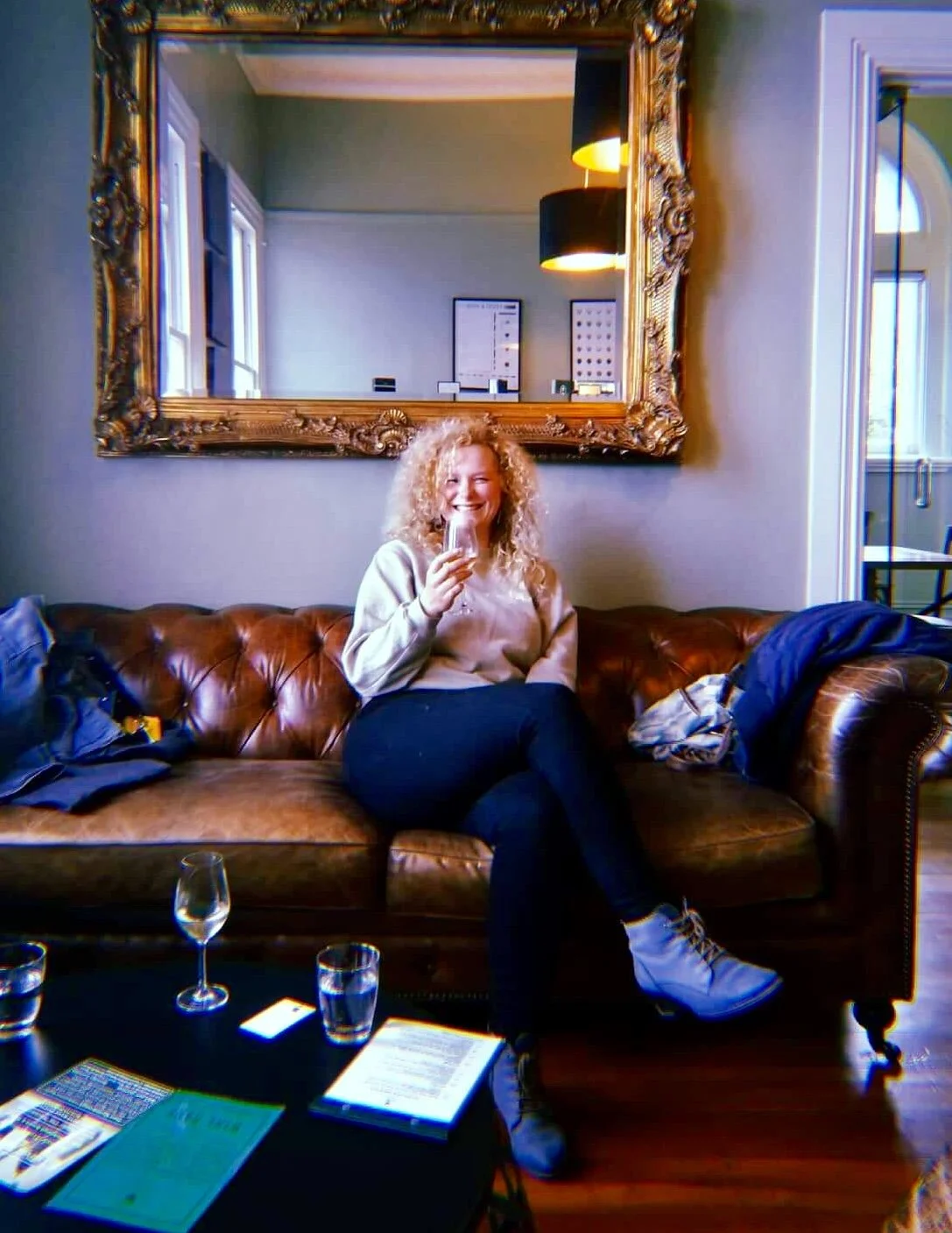 A woman with curly blonde hair sitting on a brown leather tufted sofa, holding a glass and smiling. There are glasses and papers on a black table in front of her, and a large ornate mirror on the wall behind her.