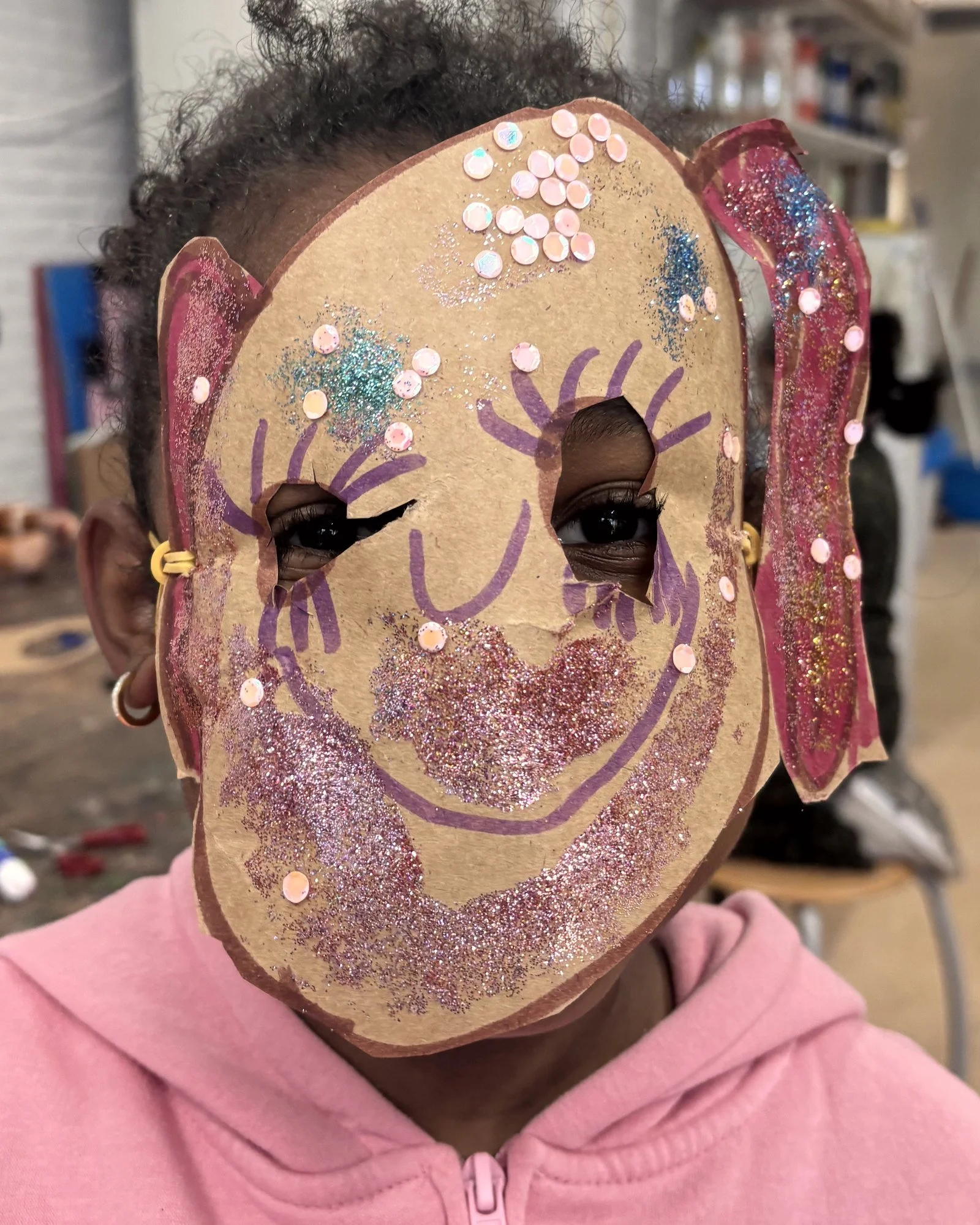 Kind houdt een masker vast dat op het gezicht lijkt, met glitters en roze pailletten, terwijl ze een roze jas draagt.