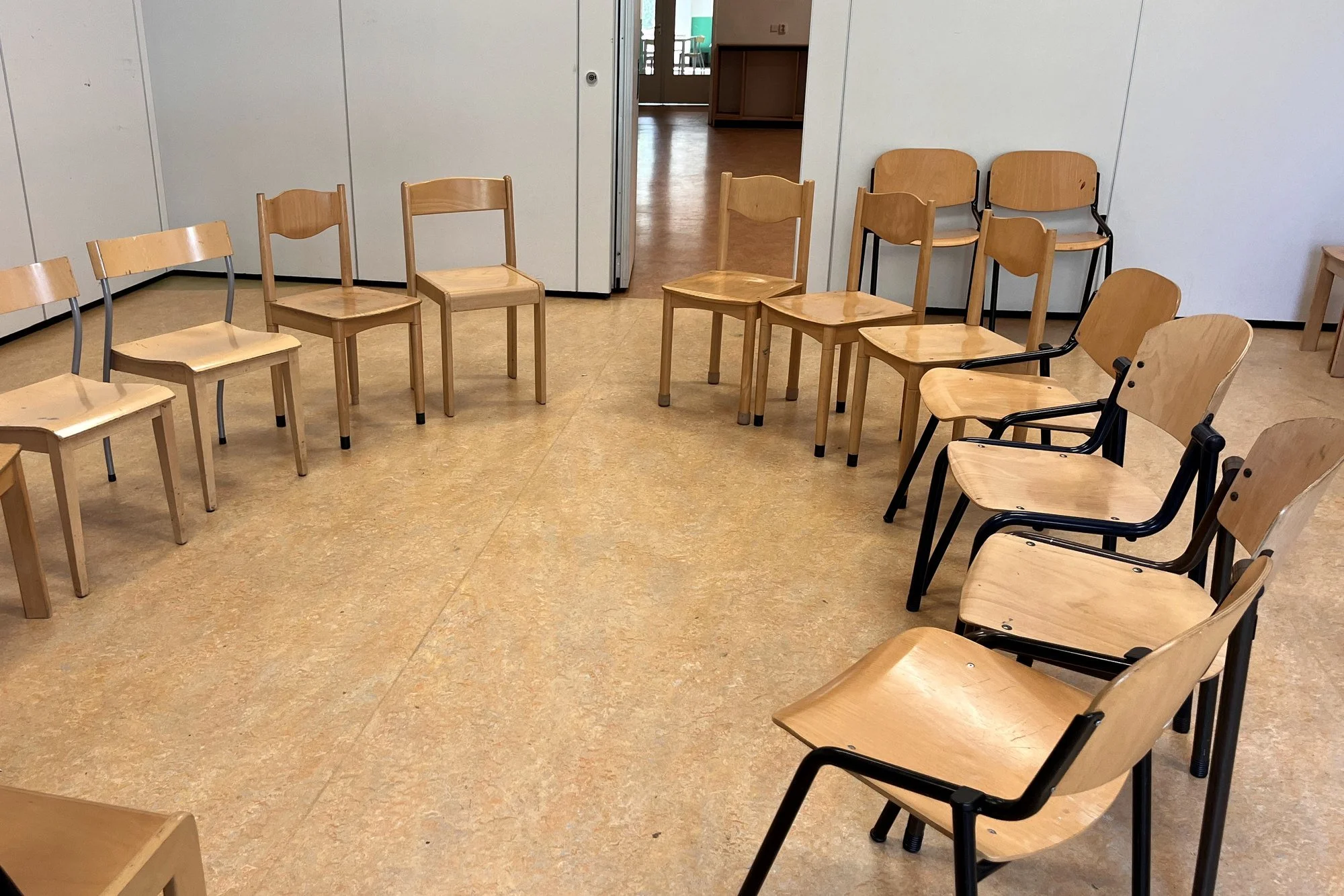 Een lege kamer met houten en metalen stoelen, enkele tegen een witte muur en andere in een cirkel, met open deur naar een andere kamer.