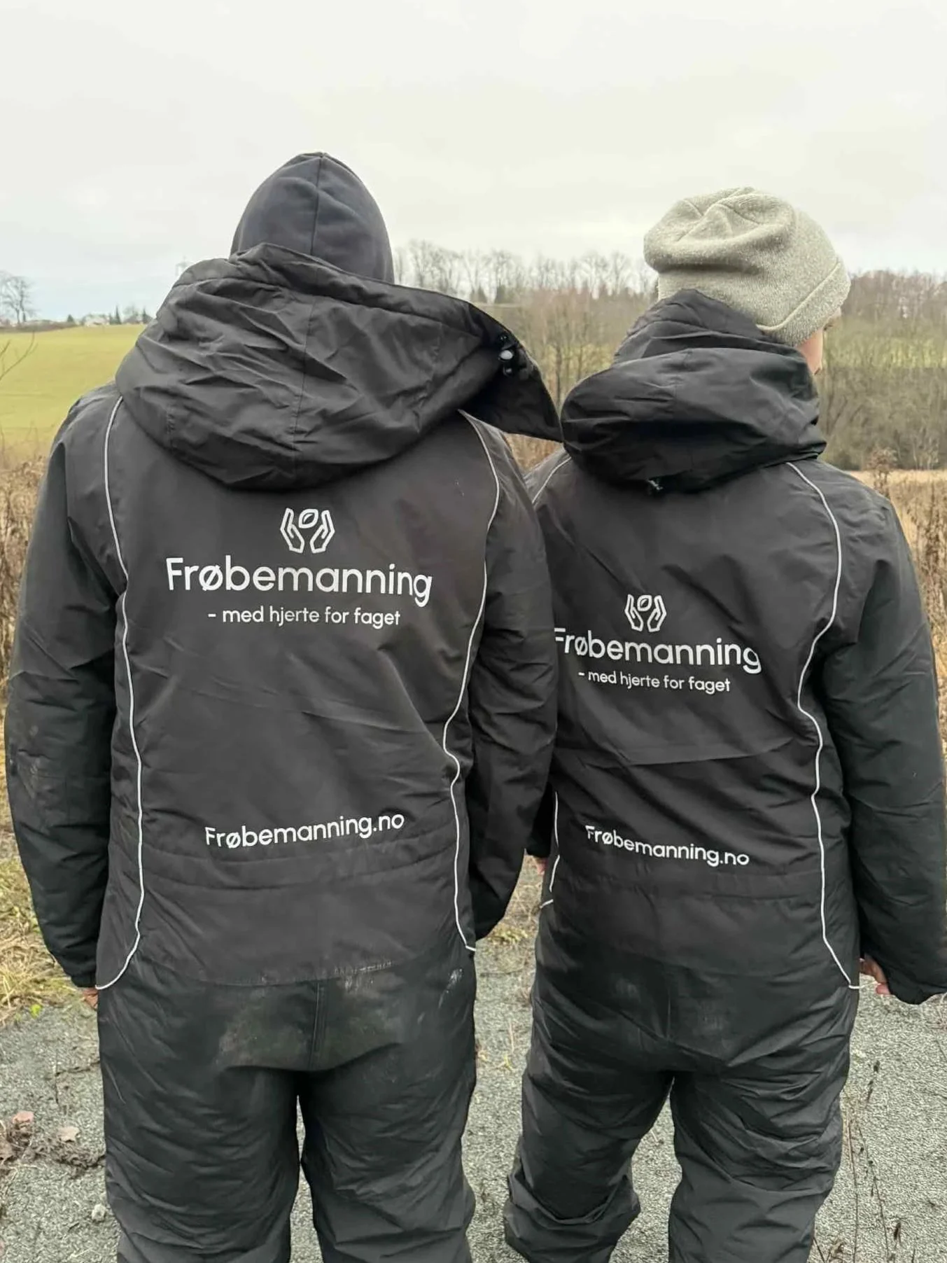 To personer fra frøbemaning står med ryggen mot kamera, iført svarte frøbemaningjakker med teksten 'Frøbemaning - med hjerte for faget' og nettstedet frøbemaning.no, utendørs med åpent landskap og trær i bakgrunnen.