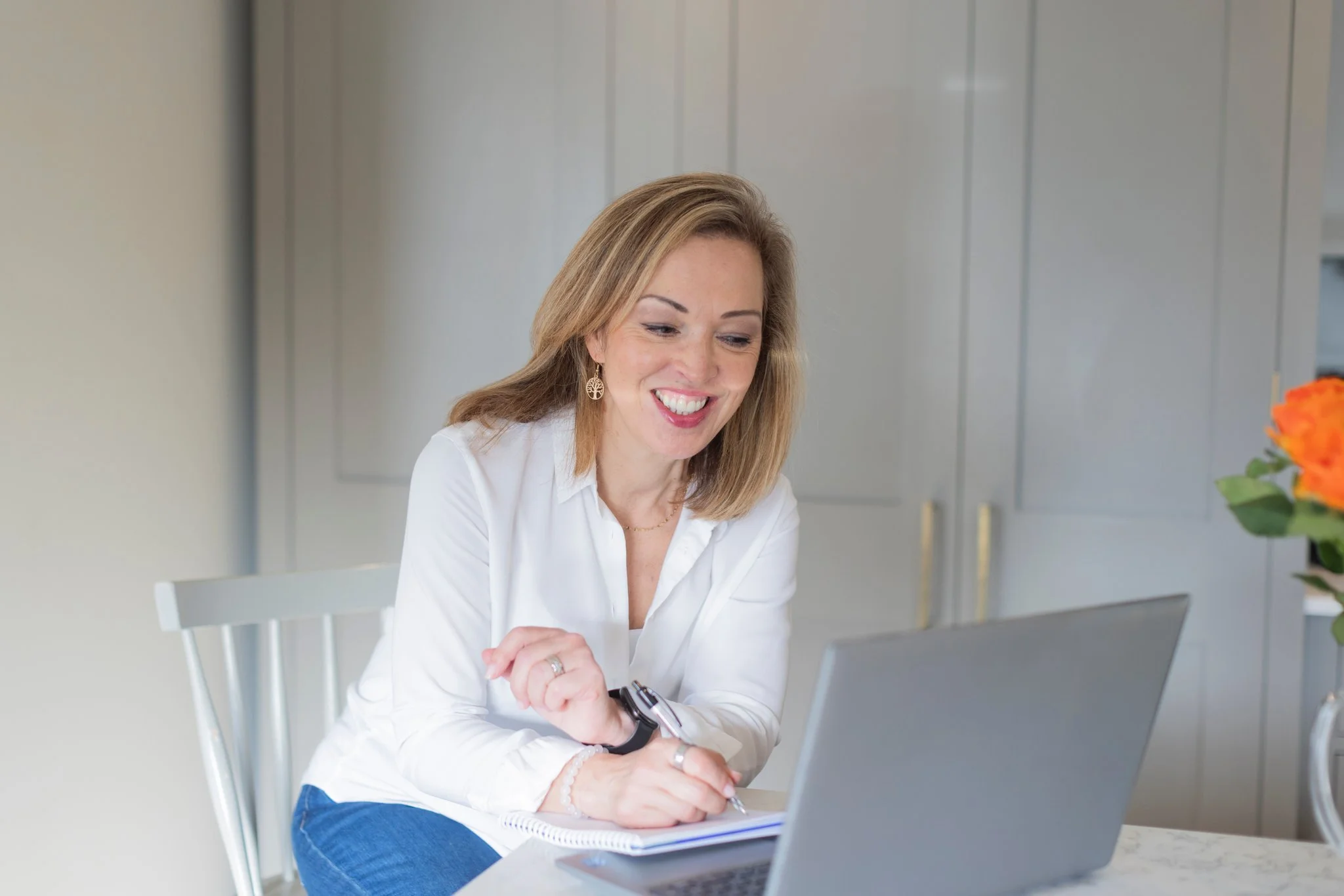 A woman with shoulder-length light brown hair, wearing a white blouse and earrings, smiling while looking at her laptop screen in a kitchen with white cabinets and a marble countertop, with a vase of orange flowers on the counter.