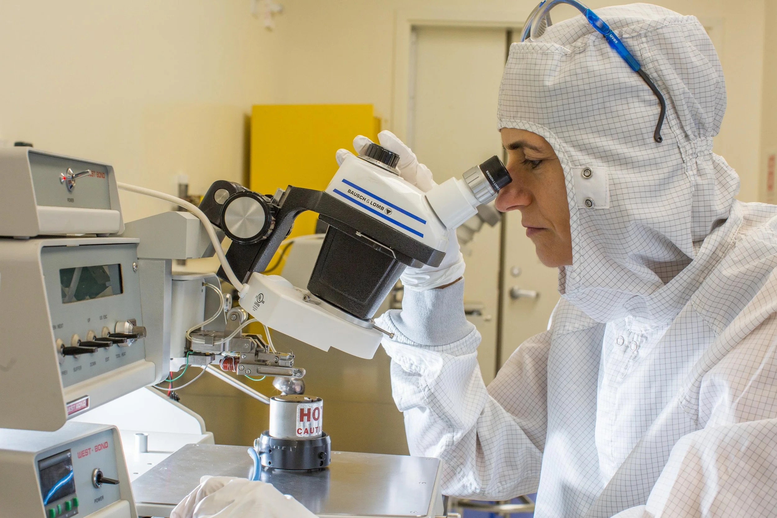 Cleanroom user operating the wire bonder, looking through its microscope to align fine wires for microelectronic device packaging.