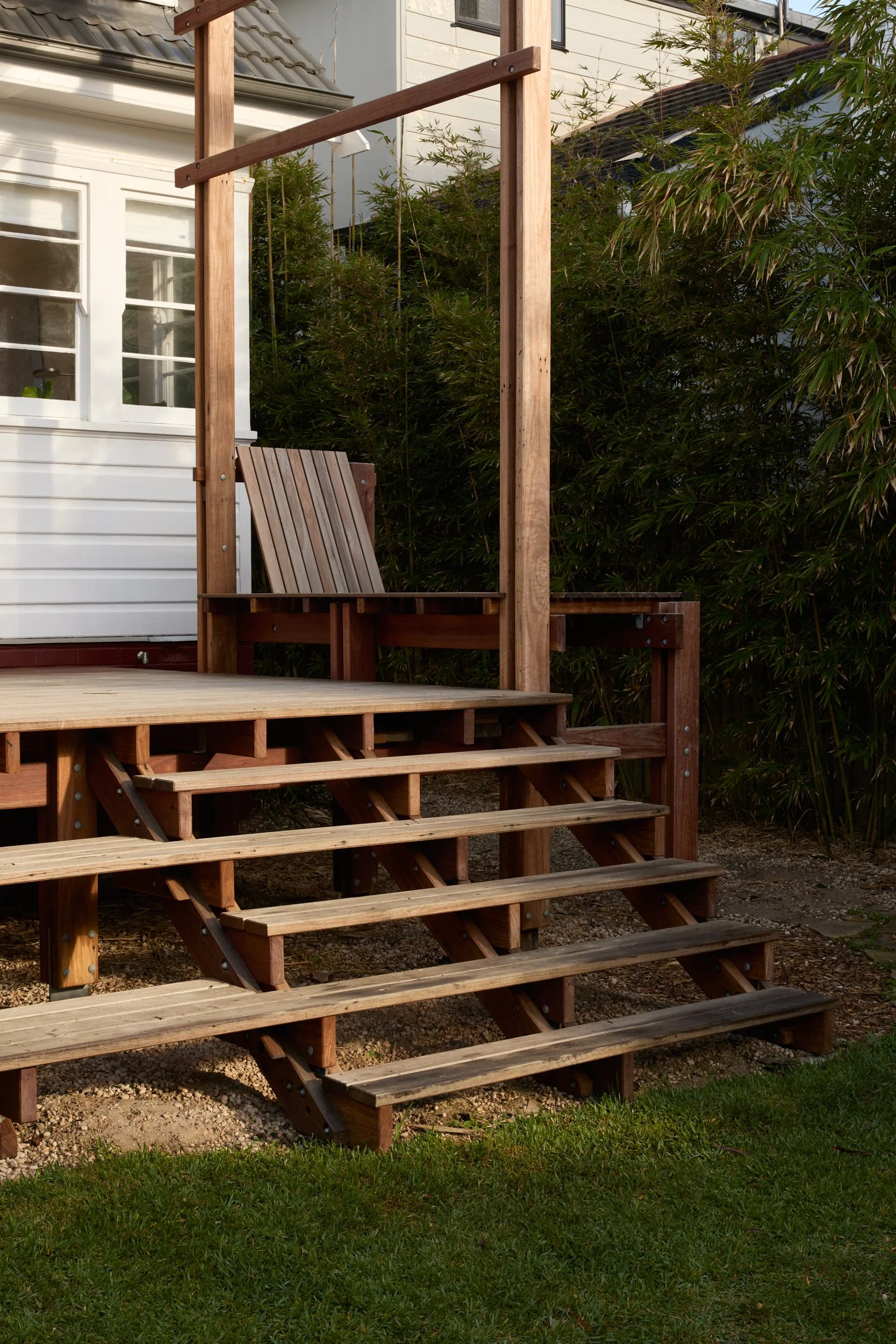 Wooden backyard deck with stairs and a swing seat, surrounded by green shrubs and a white house with window panes.