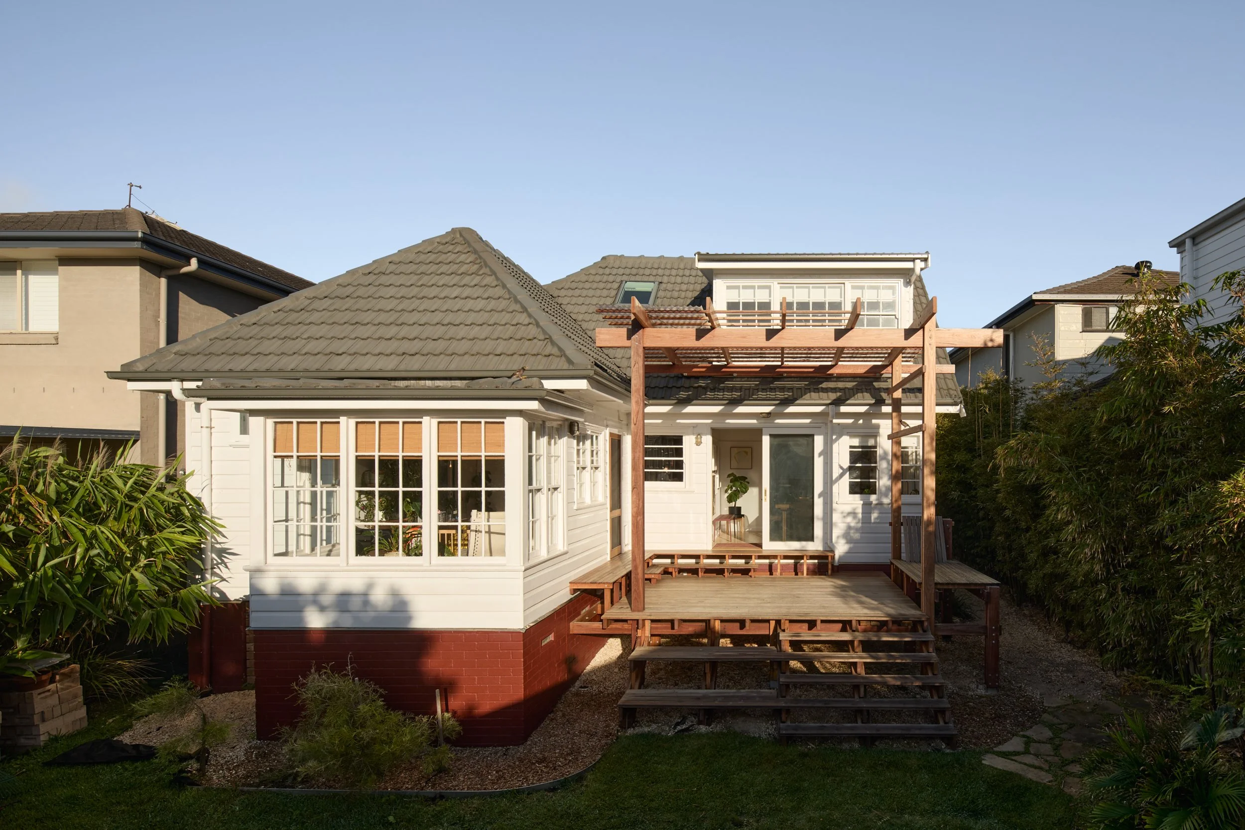 Backyard of a house with a new wooden deck under construction, surrounded by green foliage and neighboring houses with gray roofs under a clear blue sky.