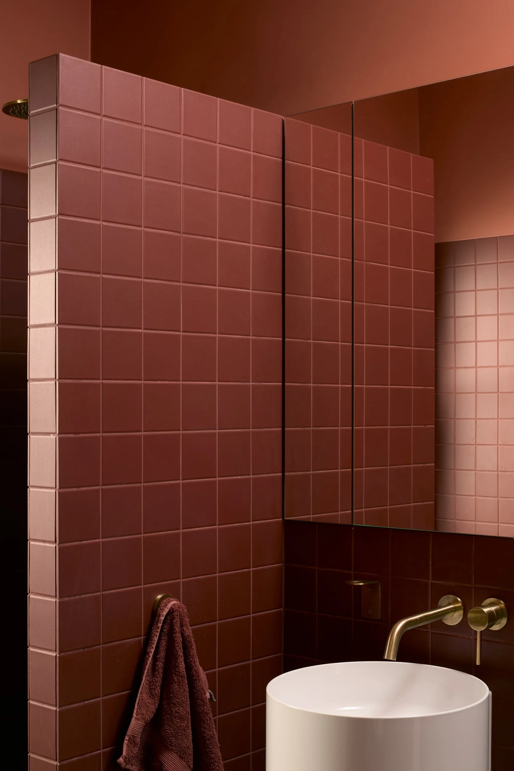 Modern bathroom with burgundy tiles, a large round white bathtub, a gold faucet, and a mirror reflecting part of the wall. A brown towel hangs on a hook on the tiled wall.