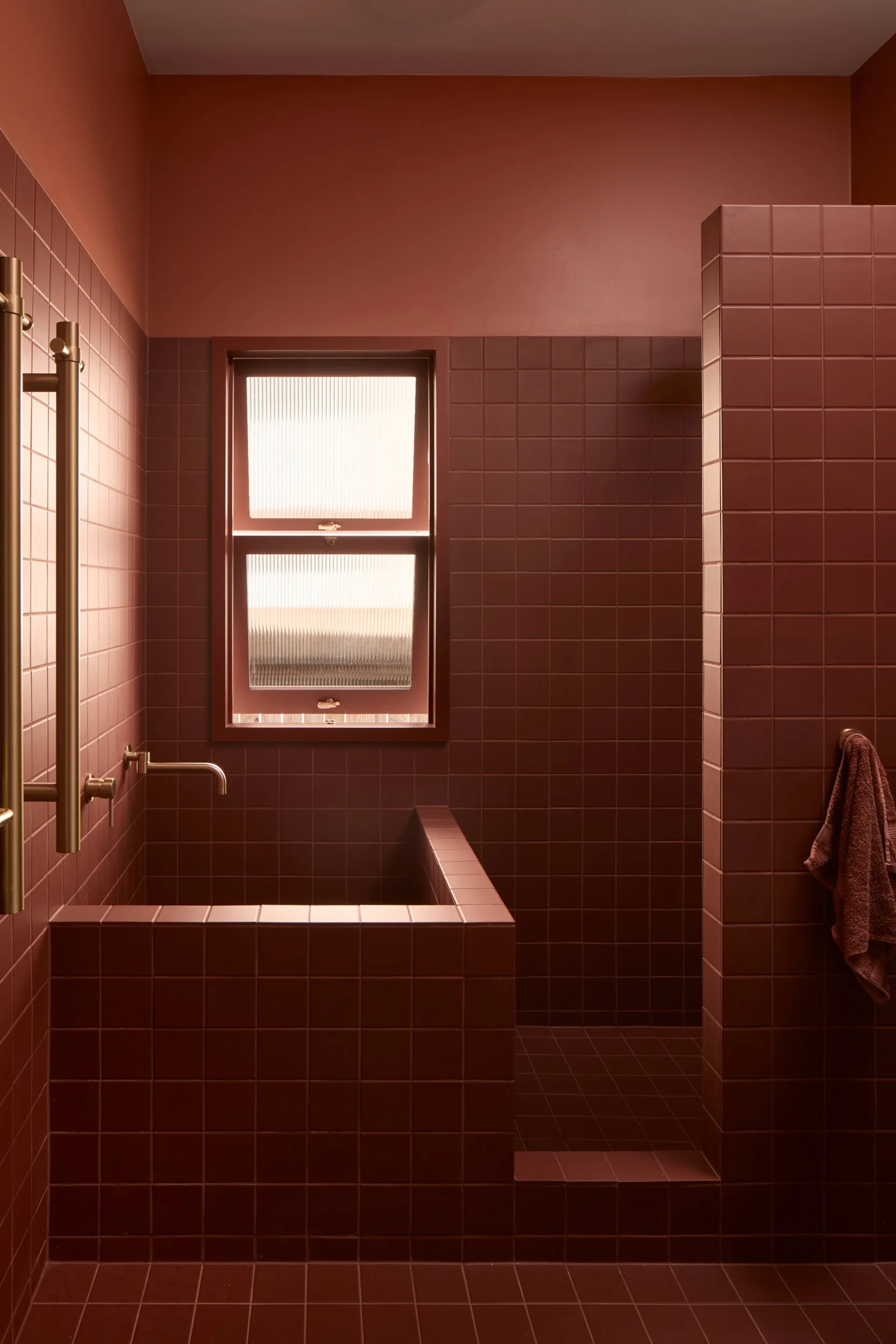 A bathroom with brown tiles on the walls and floor, a window with frosted glass, a corner shower area with a small step, and a towel hanging on a rail.