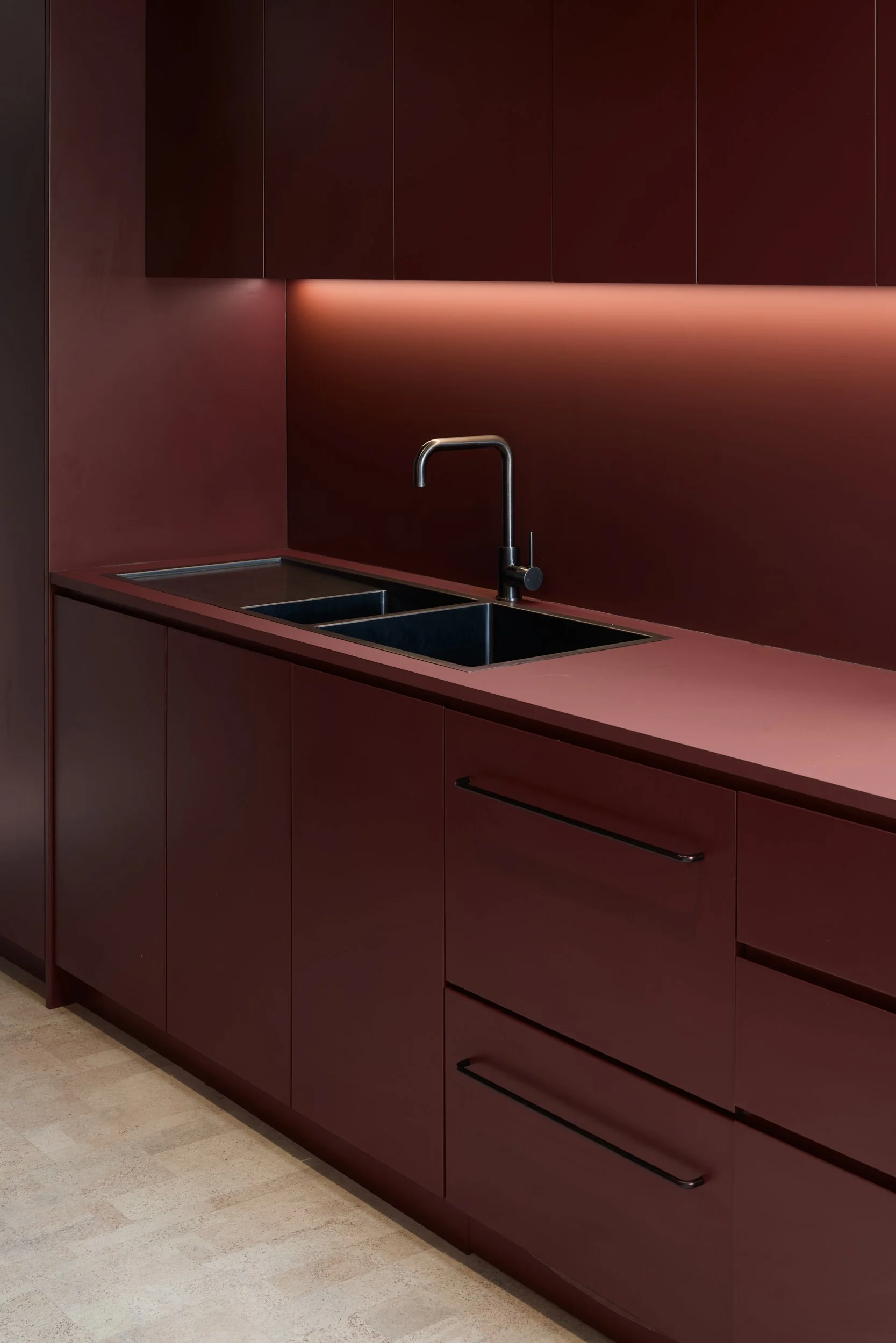 Close-up of a modern kitchen with burgundy cabinets, a black countertop, a stainless steel sink, and a black faucet, illuminated by warm under-cabinet lighting.
