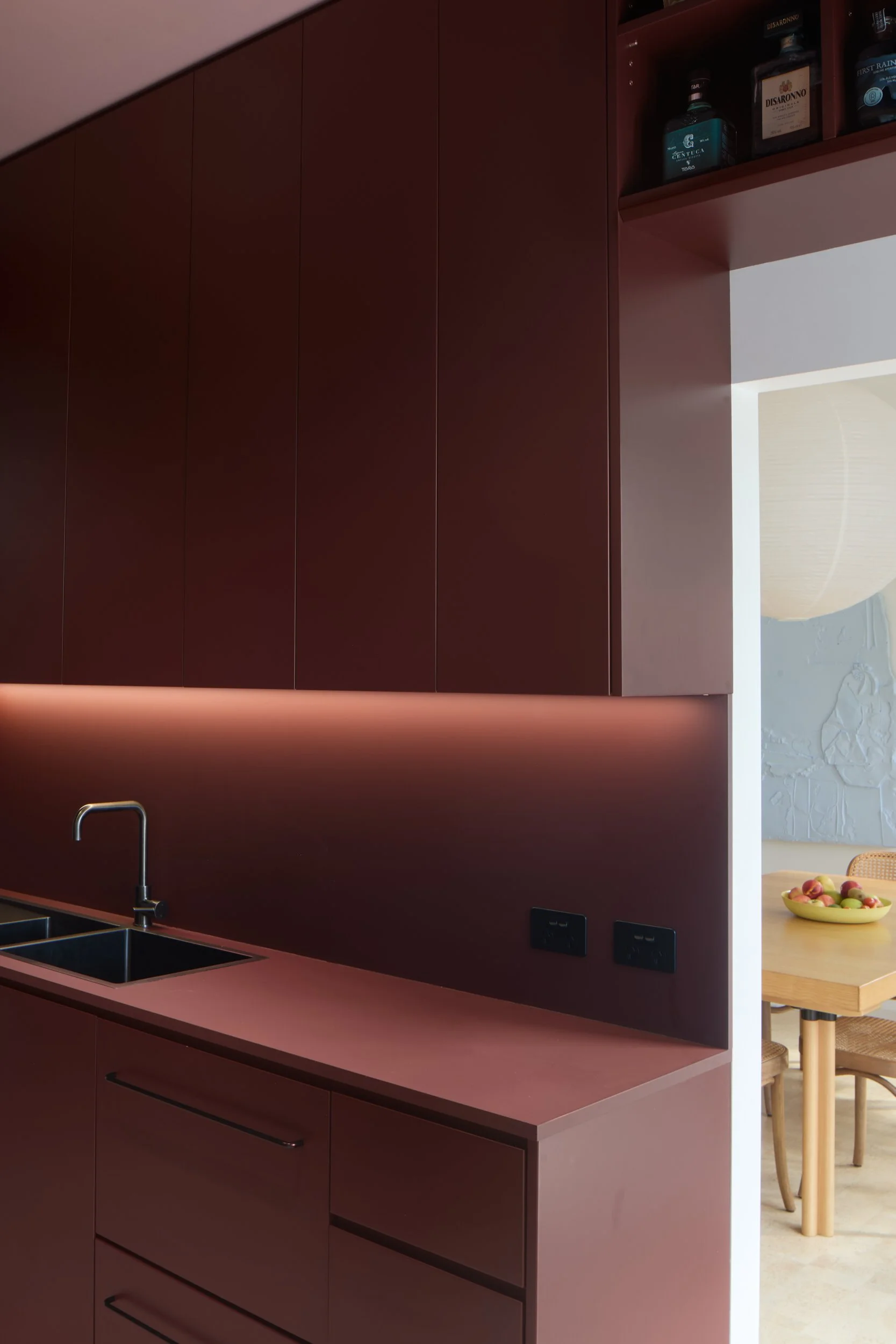 Modern kitchen with dark red cabinets, a built-in wine rack, a sink with a faucet, and a view into a dining area with a table and chairs, a bowl of fruit, and a textured wall.