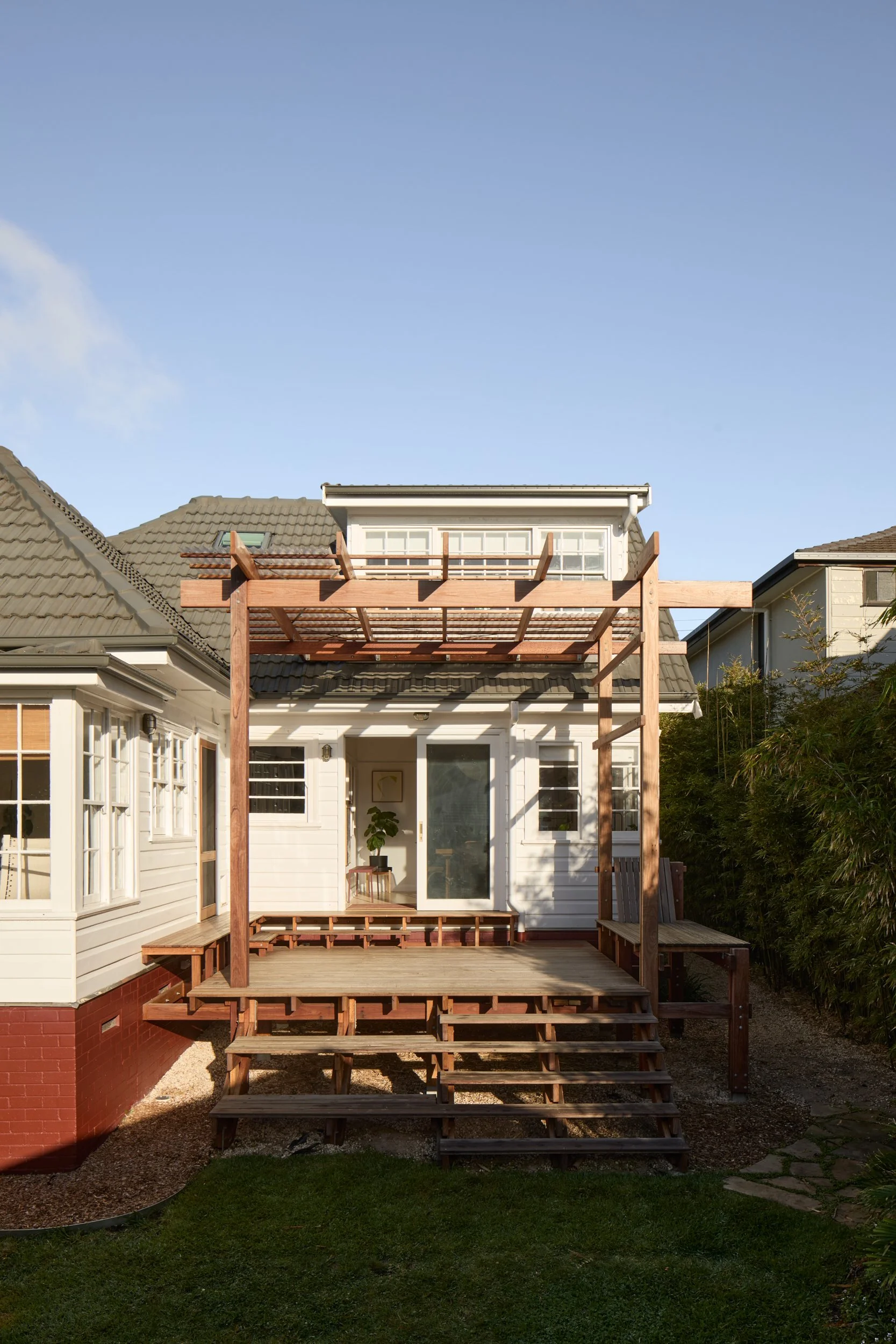 A house with a newly built wooden deck and pergola, with stairs leading up to the deck. The house has a white exterior with large windows and a sliding glass door. The background shows neighboring houses and a clear blue sky.