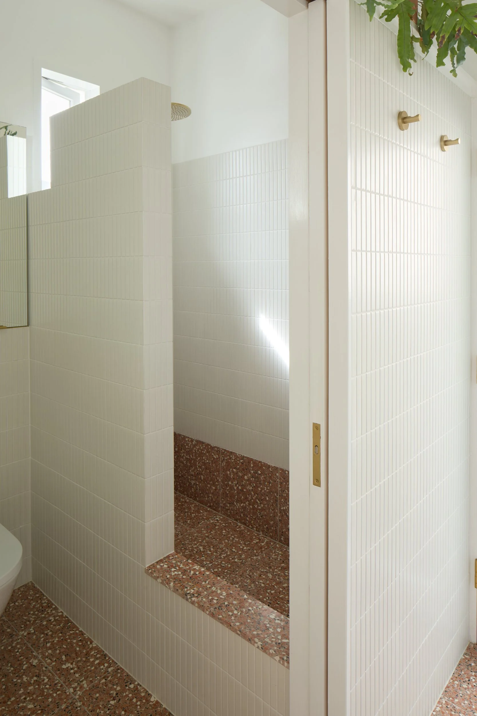 Public restroom stall with white tiled walls, a small tile shelf, a hanging plant, and a partially open door. Light coming in through a window.