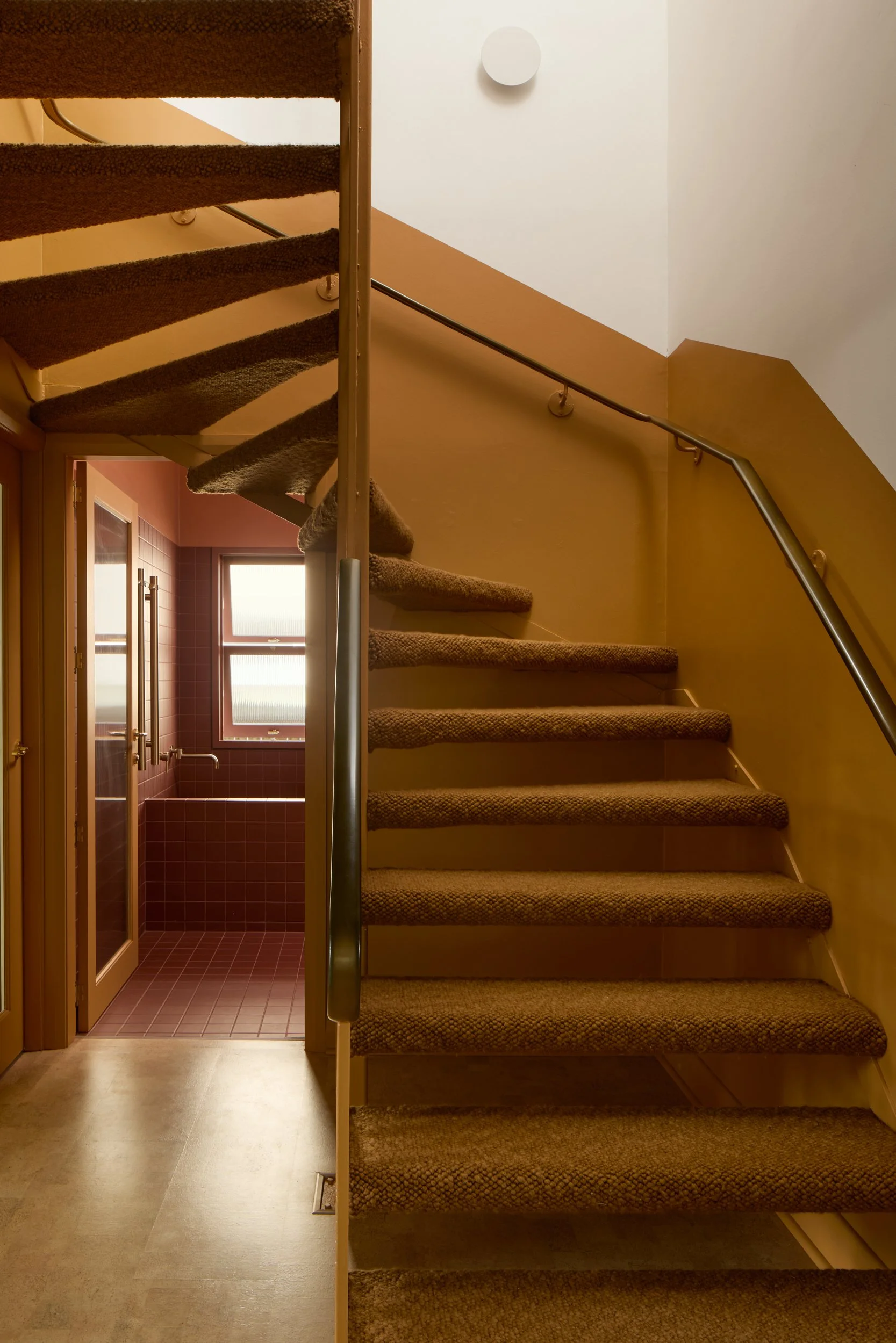 Interior view of a staircase with carpeted steps and a metal handrail, leading to a second floor with a white wall and a round wall-mounted light fixture. A small doorway with a pink-tiled bathroom is visible at the bottom of the stairs, with a windo