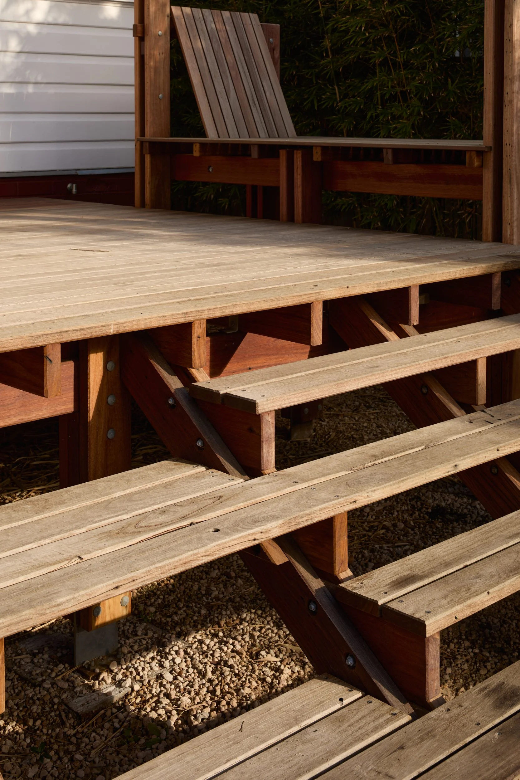 A wooden deck with steps and a built-in wooden bench, surrounded by small gravel and green shrubbery in the background.