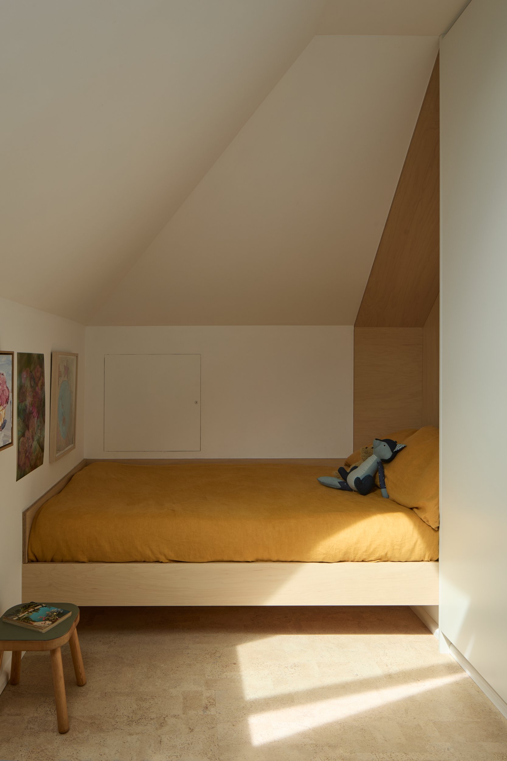 A bedroom with a sloped ceiling, a bed with a mustard yellow duvet, and a stuffed animal on the bed. There are framed pictures on the wall, a small table with a book, and sunlight casting a shadow on the floor.