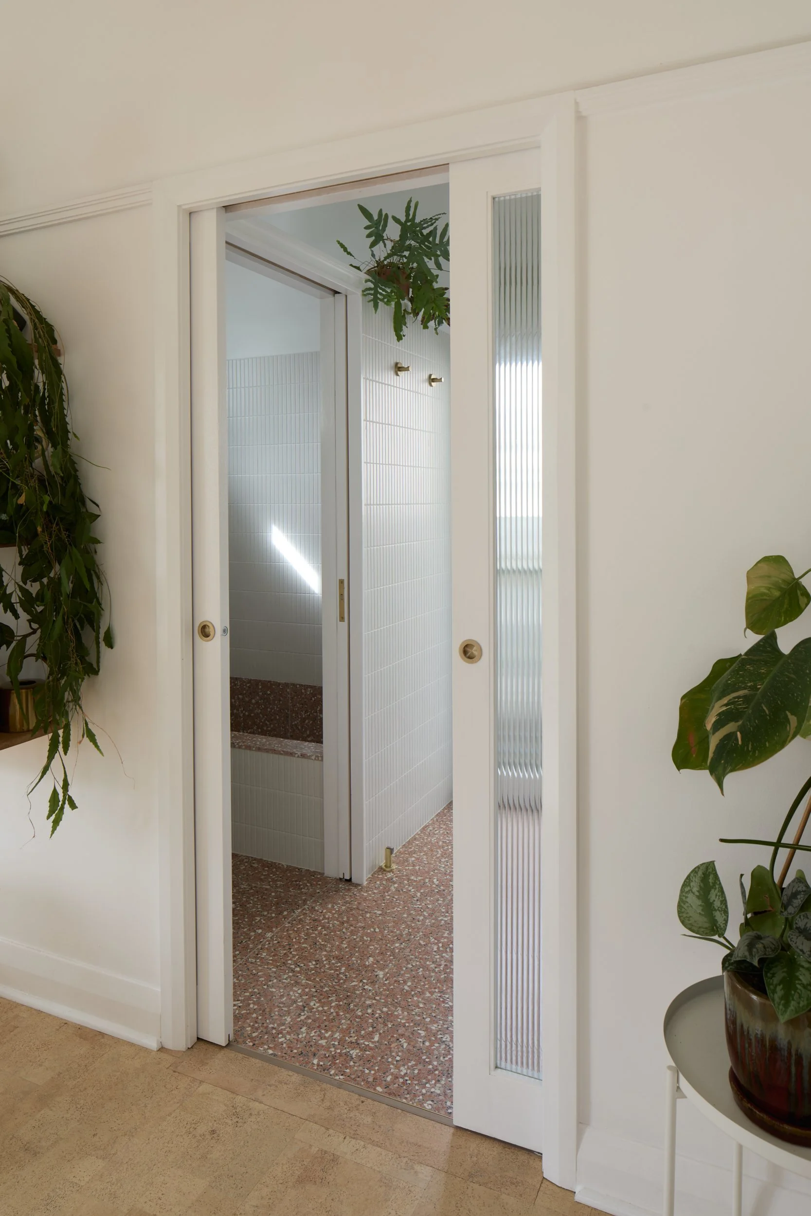 A white sliding door with frosted glass panels leading to a tiled bathroom, decorated with potted plants on either side, floor is beige tile.