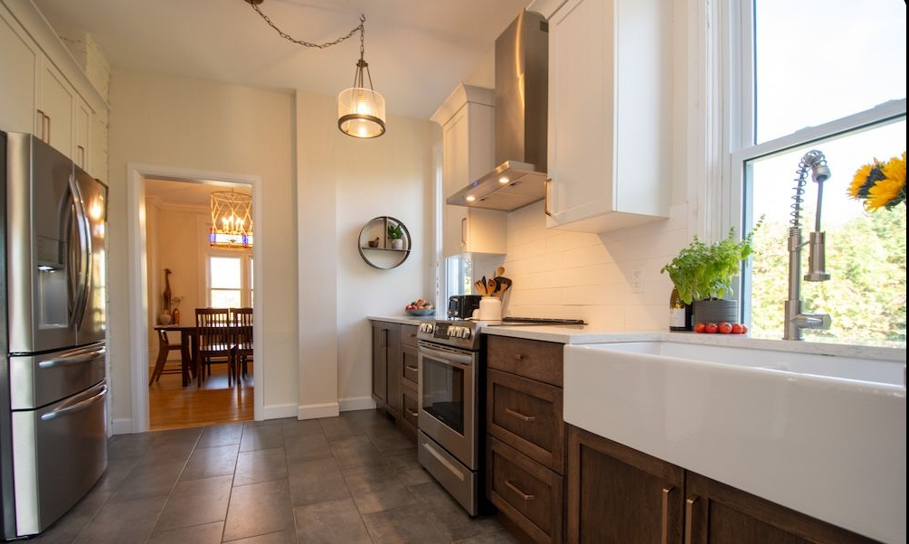 Kitchen with stainless steel refrigerator, white cabinets, dark wood lower cabinets, white farmhouse sink, stove, and a window with a view of trees, some potted plants, and cherry tomatoes on the counter.