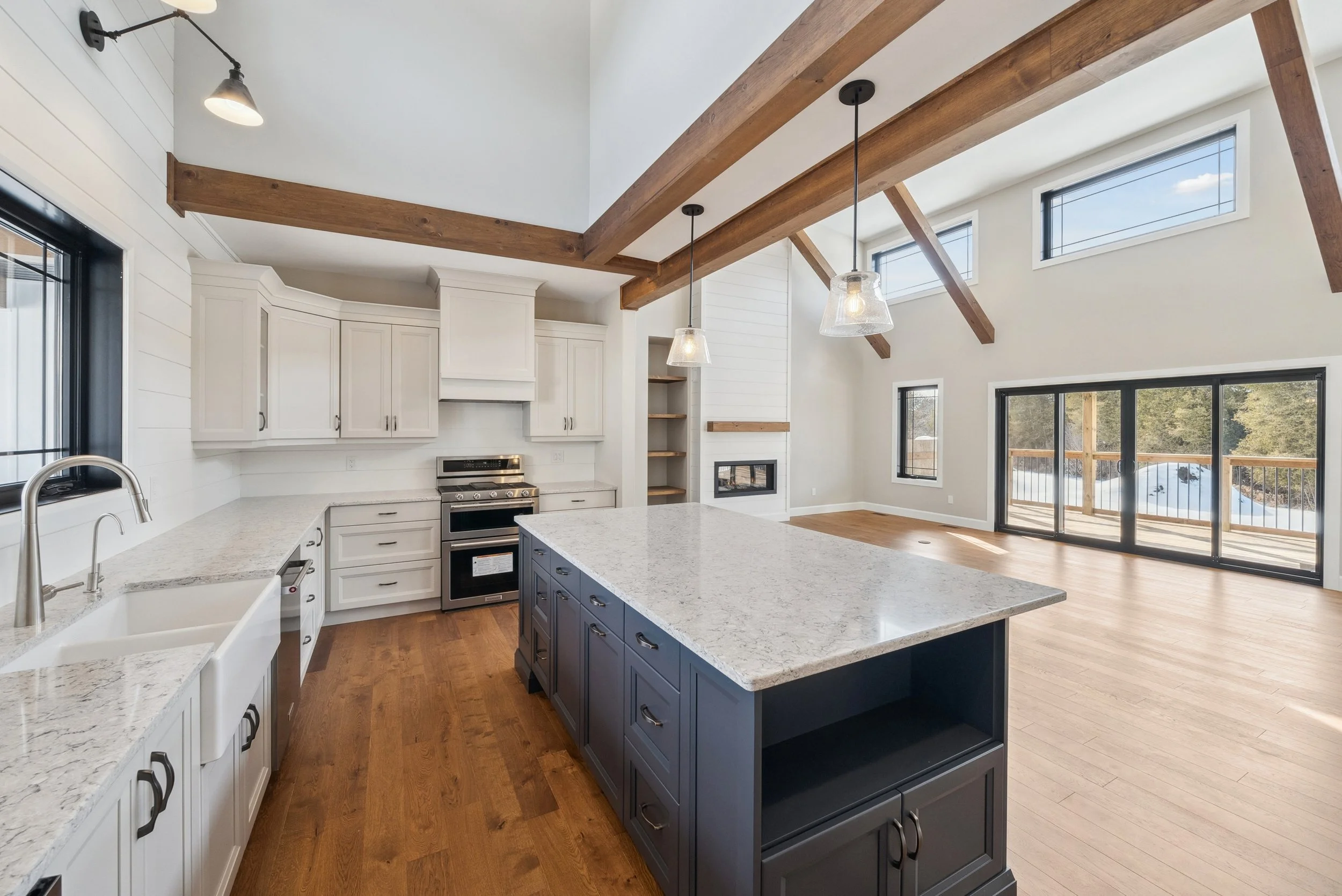 Open-concept kitchen with white cabinets, a navy blue island, granite countertops, hardwood floors, high vaulted ceilings with exposed wooden beams, large windows, and sliding glass doors leading to a deck.