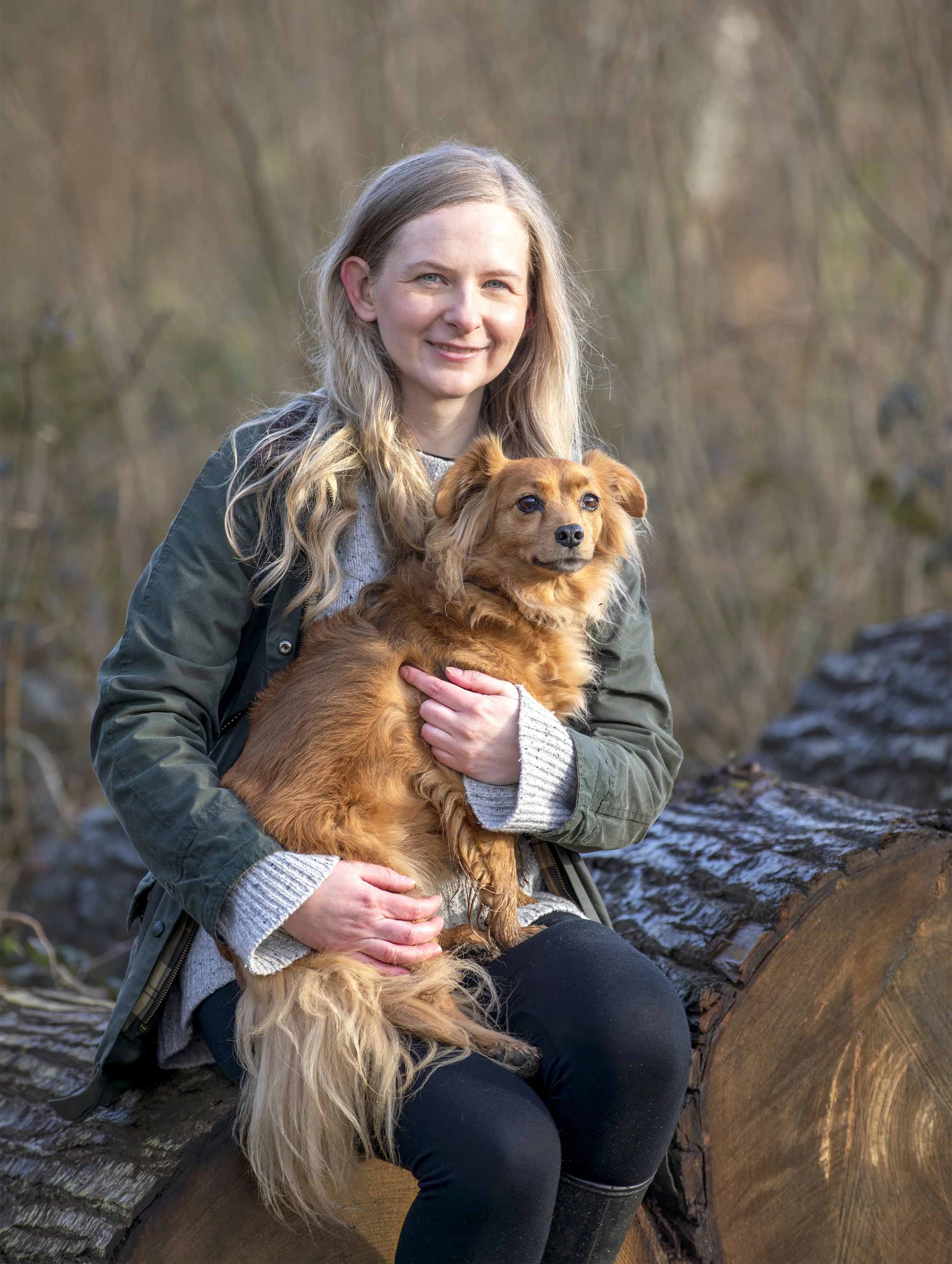 Smiling woman with blonde hair sitting on a tree log holding a small golden dog in woodland