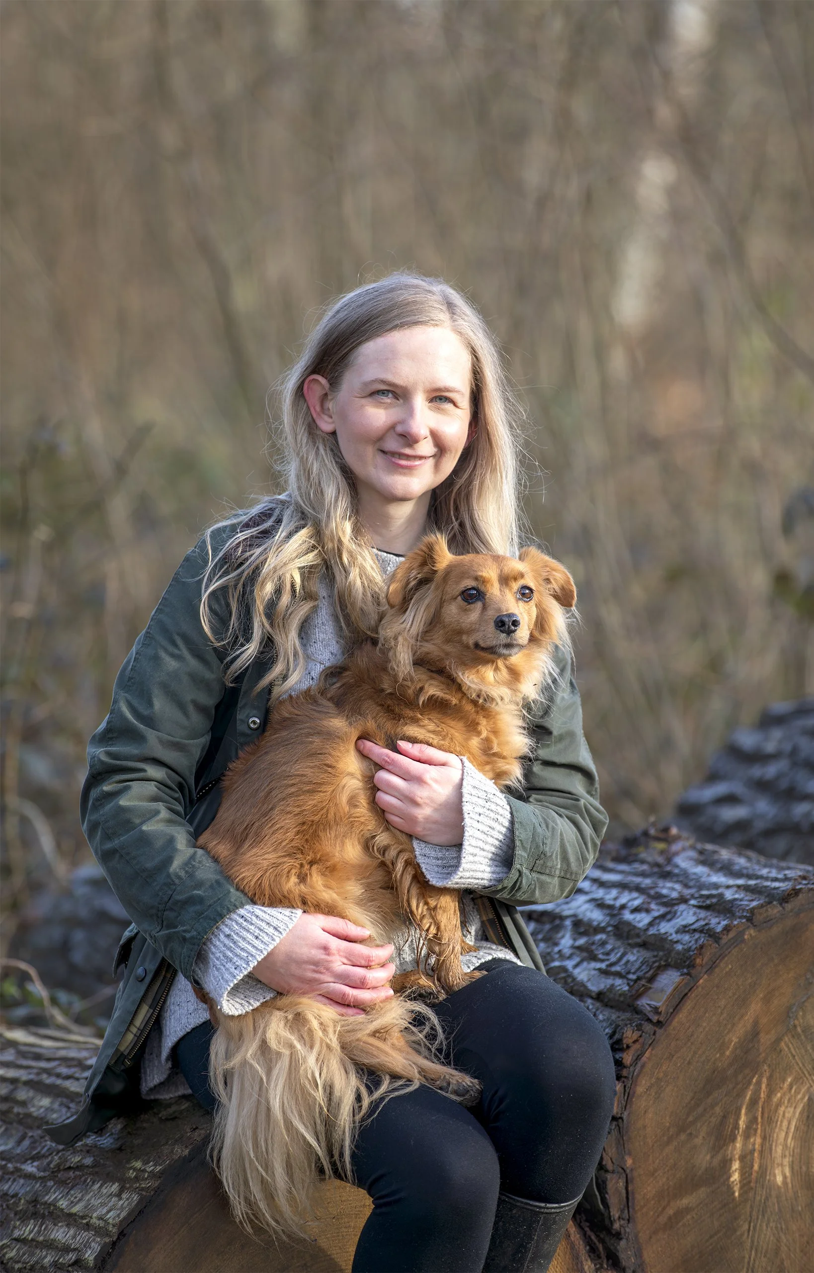 Smiling woman with blonde hair sitting on a tree log holding a small golden dog in woodland