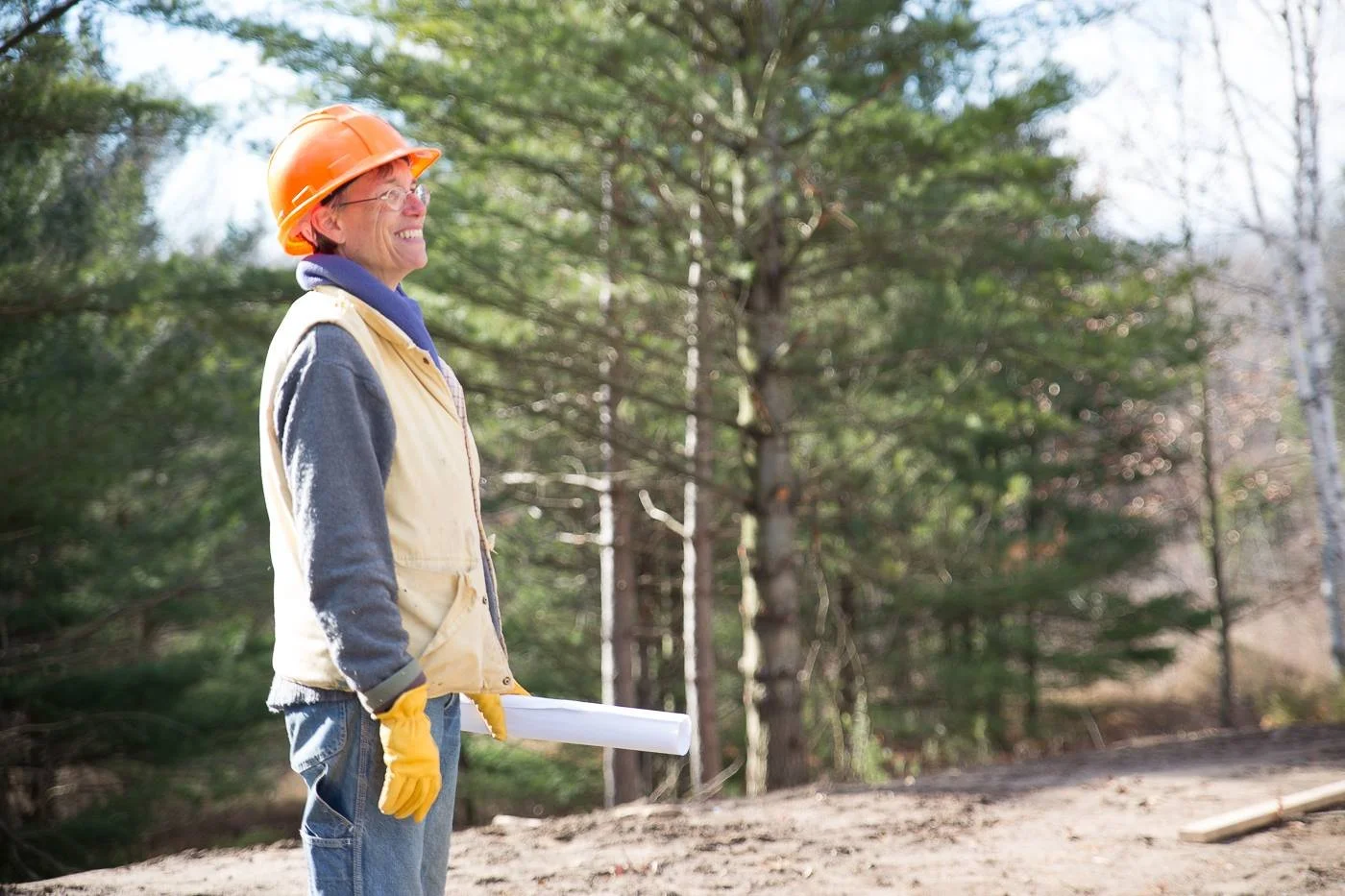 A smiling woman wearing a yellow safety vest, orange hard hat, yellow gloves, and glasses stands outdoors in a wooded area, holding rolled-up papers.