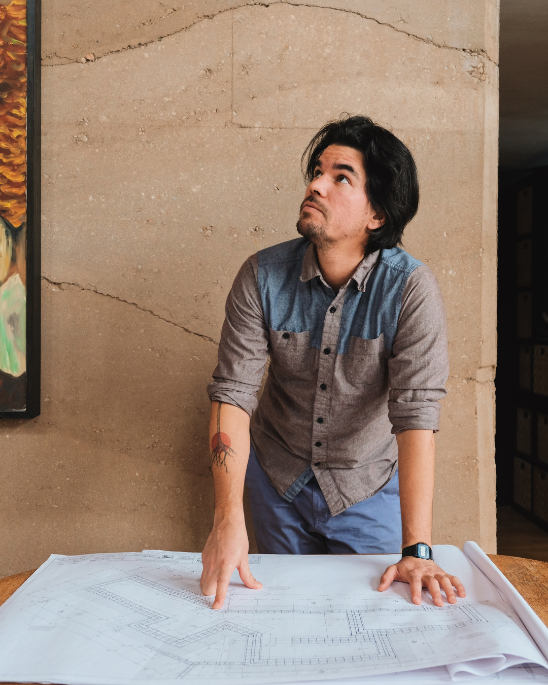 A man with black hair and a tattoo on his left arm is leaning over a table with blueprints, looking up thoughtfully.