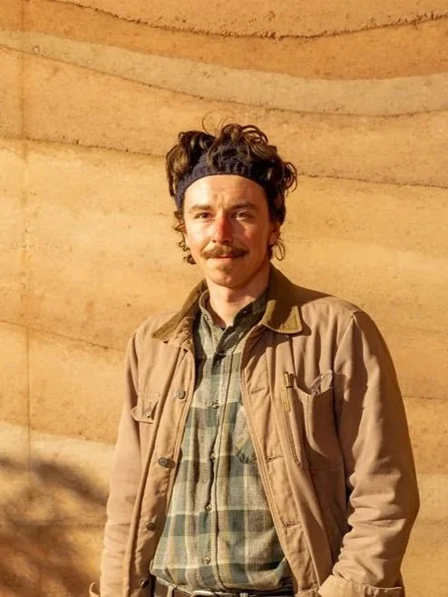 A man with curly hair, a mustache, and a headband standing in front of a rammed earth wall.wearing a light brown jacket and plaid shirt.