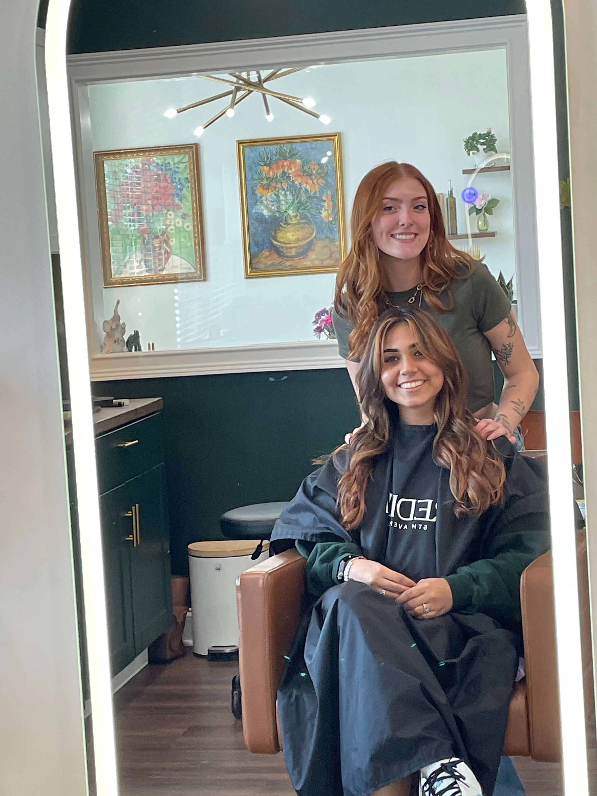A woman sitting in a salon chair with a Master Cosmetologist Annamarie Watkins standing behind her, smiling, inside a 200 East Mane Salon with artwork on the wall and a modern light fixture.