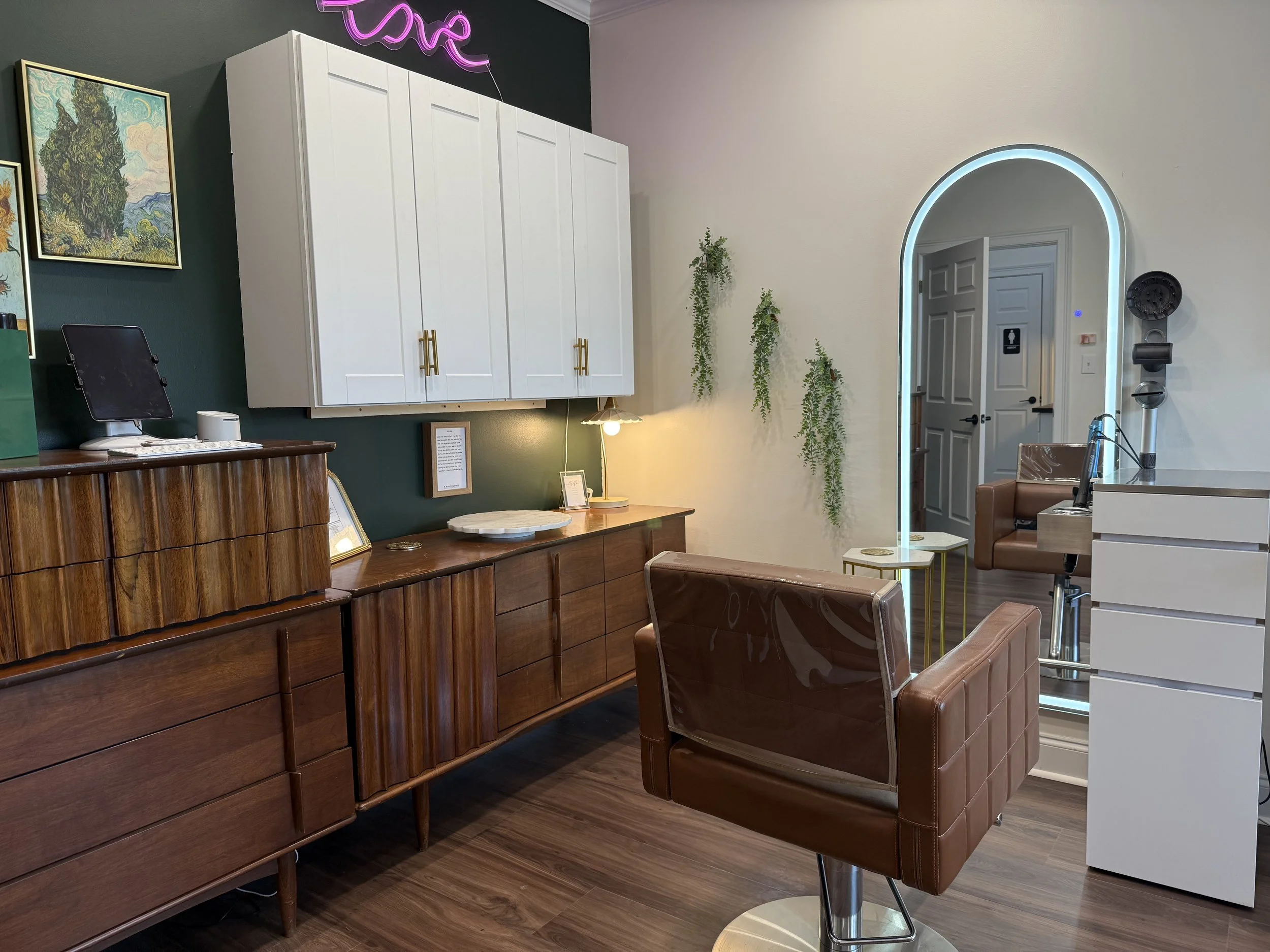 Inside Suzi Loves Hair suite, with wooden reception desk, a brown salon chair with a plastic cover, white cabinets, a mirror with an LED light, a small side table, and wall decor including hanging plants and a neon pink sign.