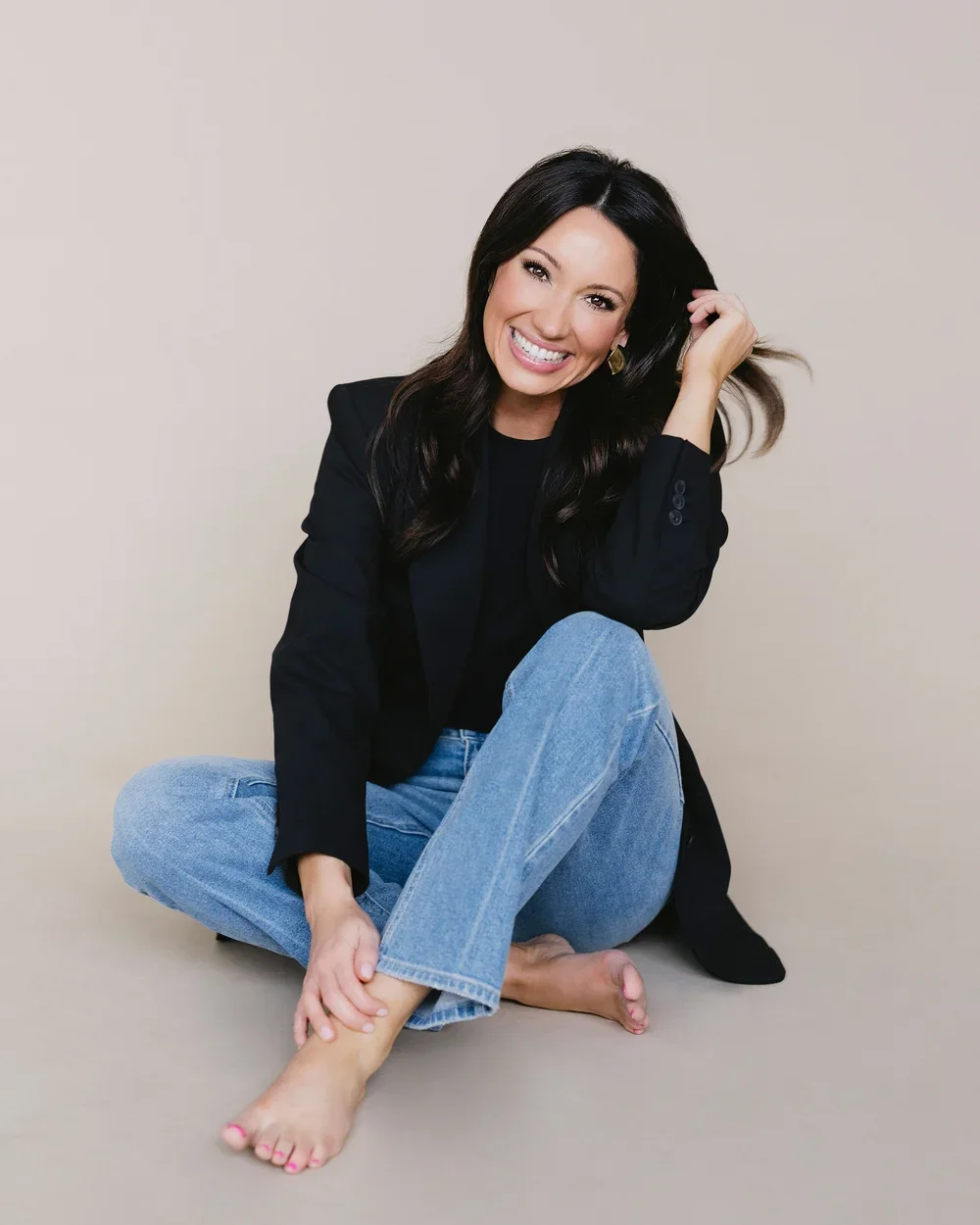A woman with long dark hair, wearing a black blazer and blue jeans, sitting barefoot on the floor against a plain beige background, smiling and touching her hair.