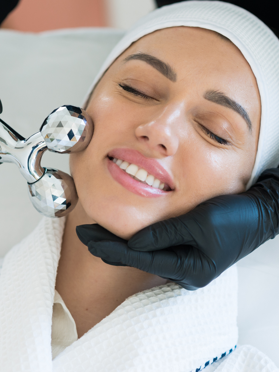 A woman receiving a facial massage with a jade roller, lying down with her eyes closed and smiling, in a spa setting.