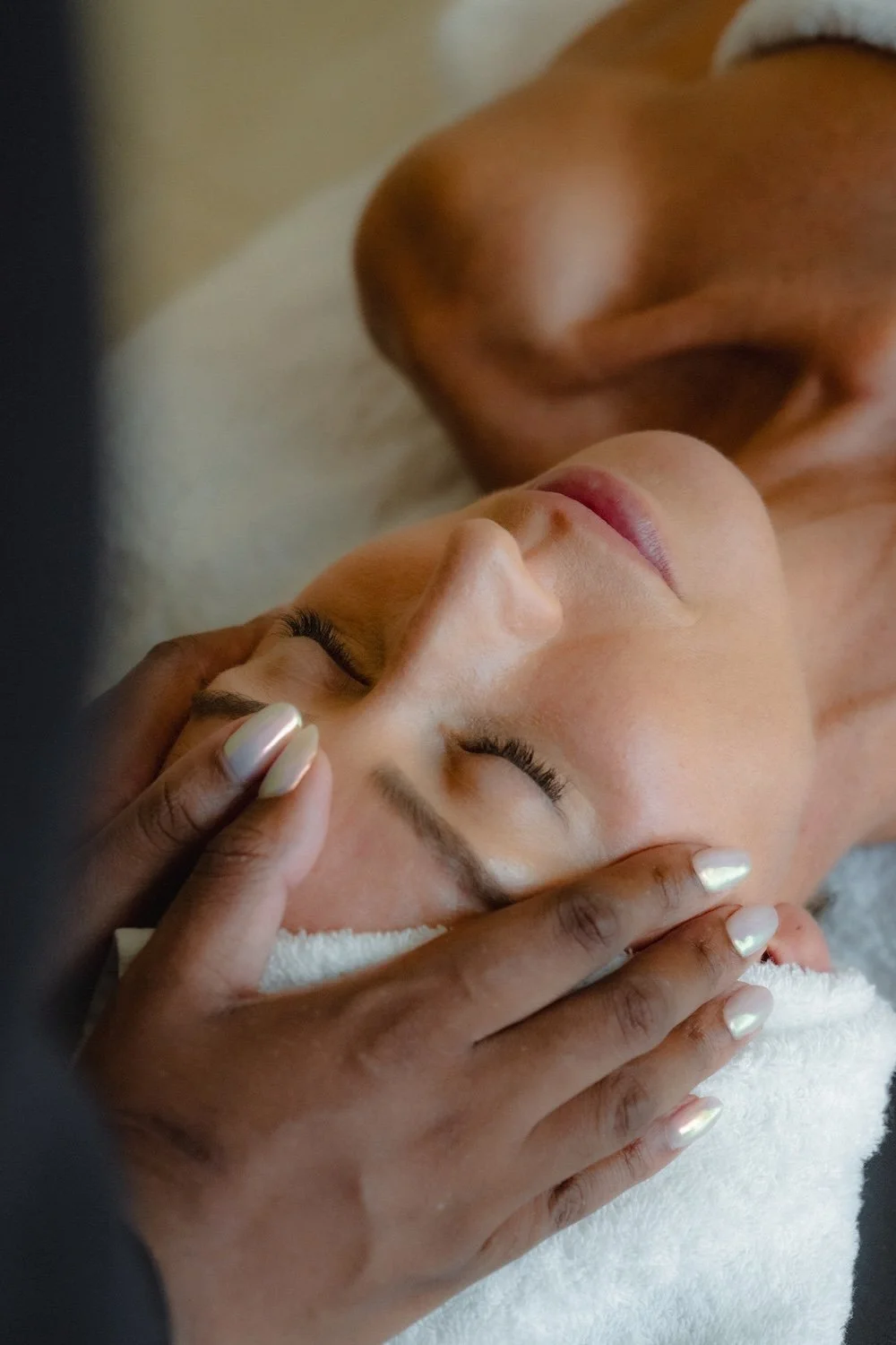 A woman receives a facial massage in a spa setting, lying on a towel with eyes closed, while a facial therapist's hands gently touch her forehead.