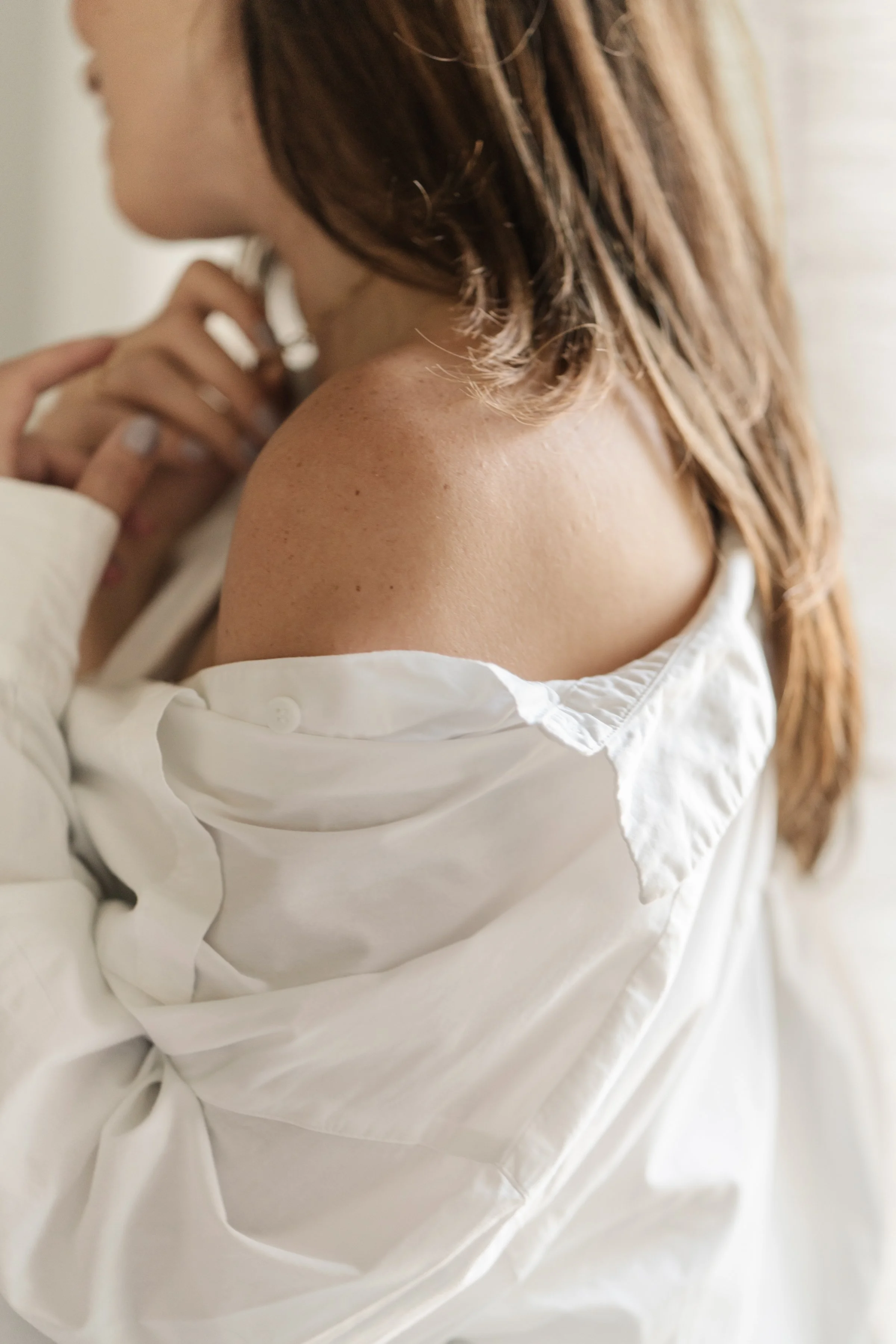 Close-up of a woman with light brown hair, wearing a white shirt with the collar visible, holding her hand near her face.