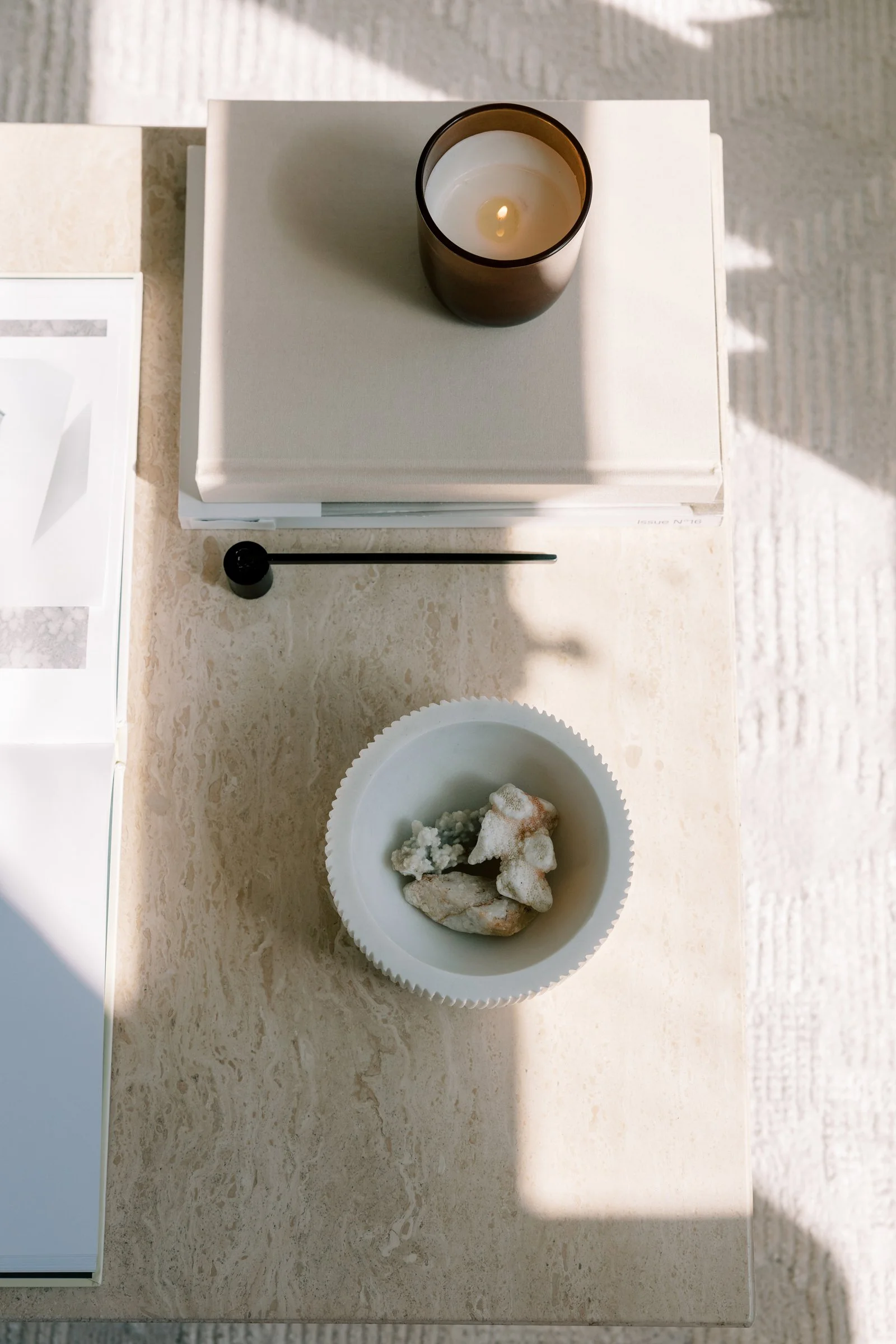 Ceramic bowl filled with irregularly shaped rocks or mineral specimens on a beige marble countertop. A candle in a glass holder is placed on a white book or box next to the bowl. A part of a white picture frame is visible on the left side.