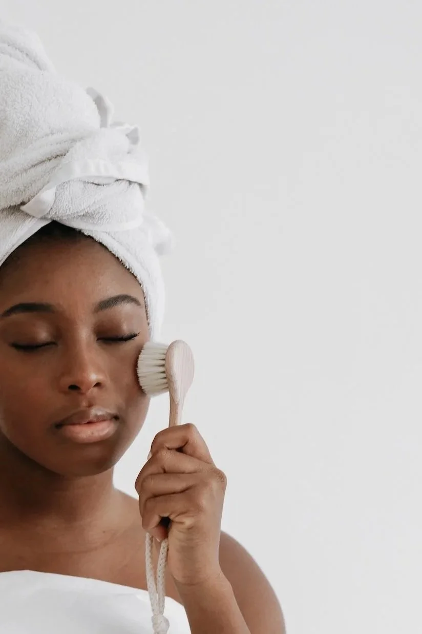 A woman with a white towel wrapped around her head gently holding a facial brush to her cheek with her eyes closed.